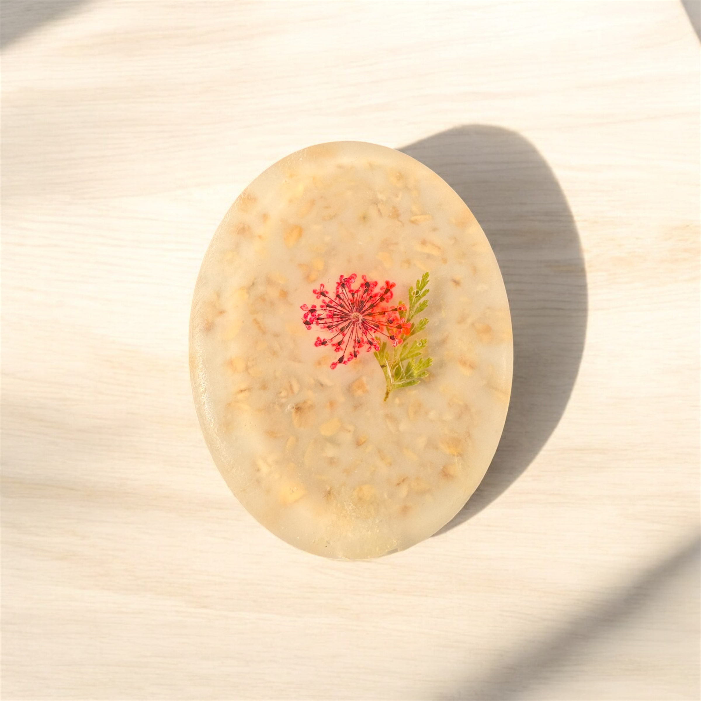 Un savon ovale avec des morceaux d'avoine et une fleur rose décorative sur le dessus, posé sur une surface en bois clair.