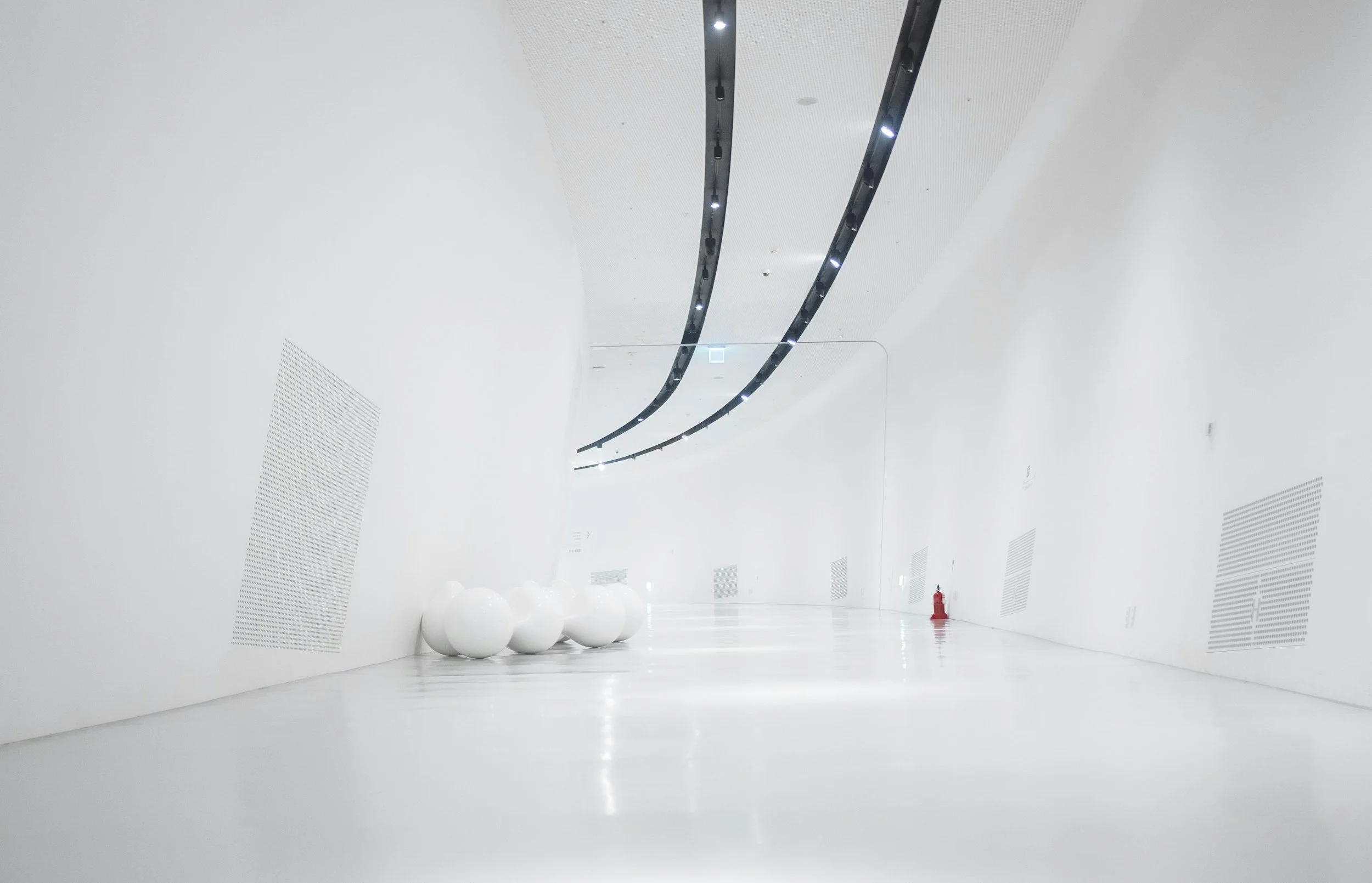 Un couloir moderne et minimaliste avec des murs blancs, un sol blanc brillant, des luminaires noirs au plafond, des objets sphériques blancs alignés contre le mur et un extincteur rouge sur le côté.