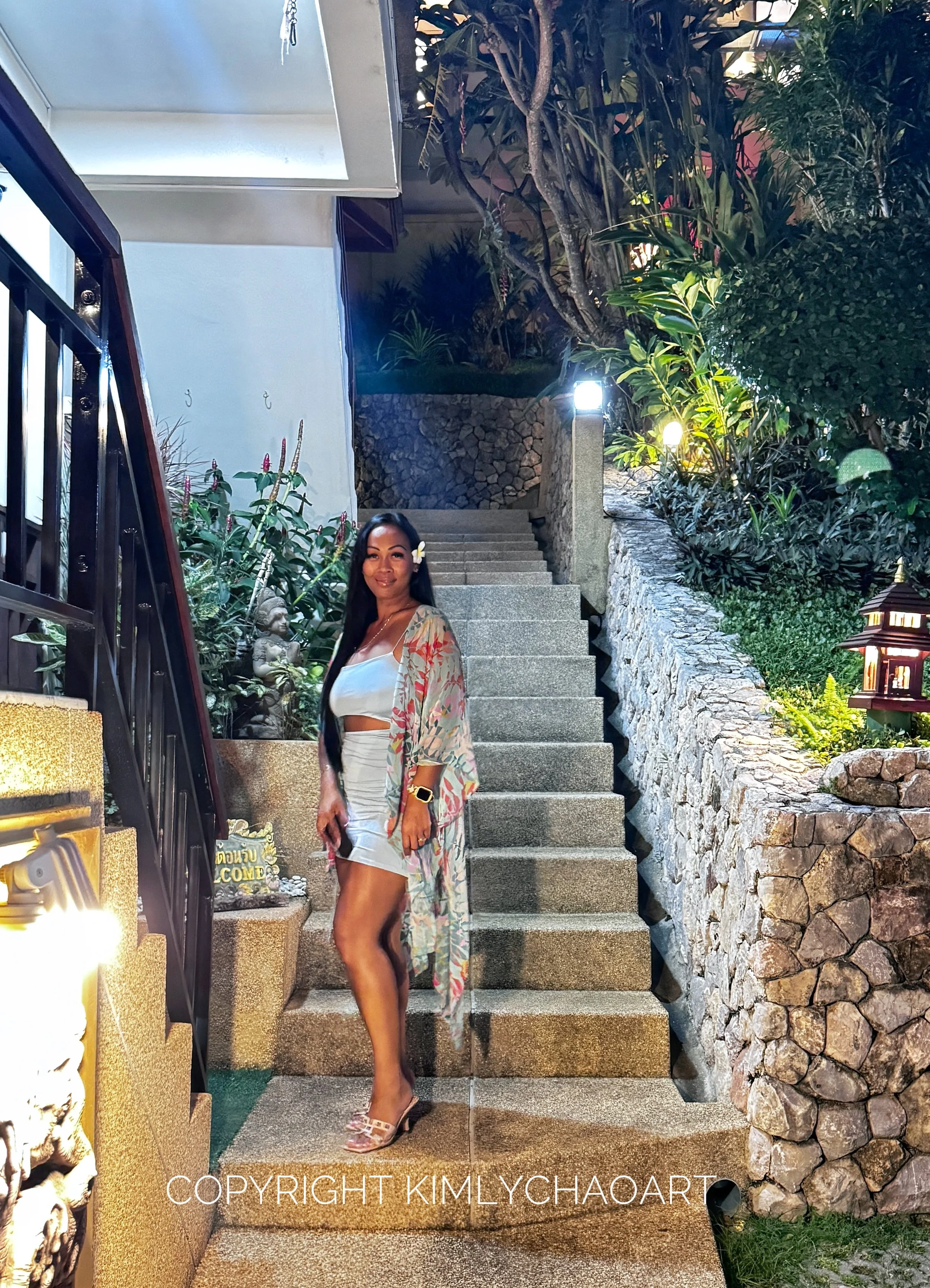 Une femme en robe blanche et kimono floral pose sur un escalier en pierre entouré de végétation et de lumières, avec une statue et un panneau de bienvenue à côté.