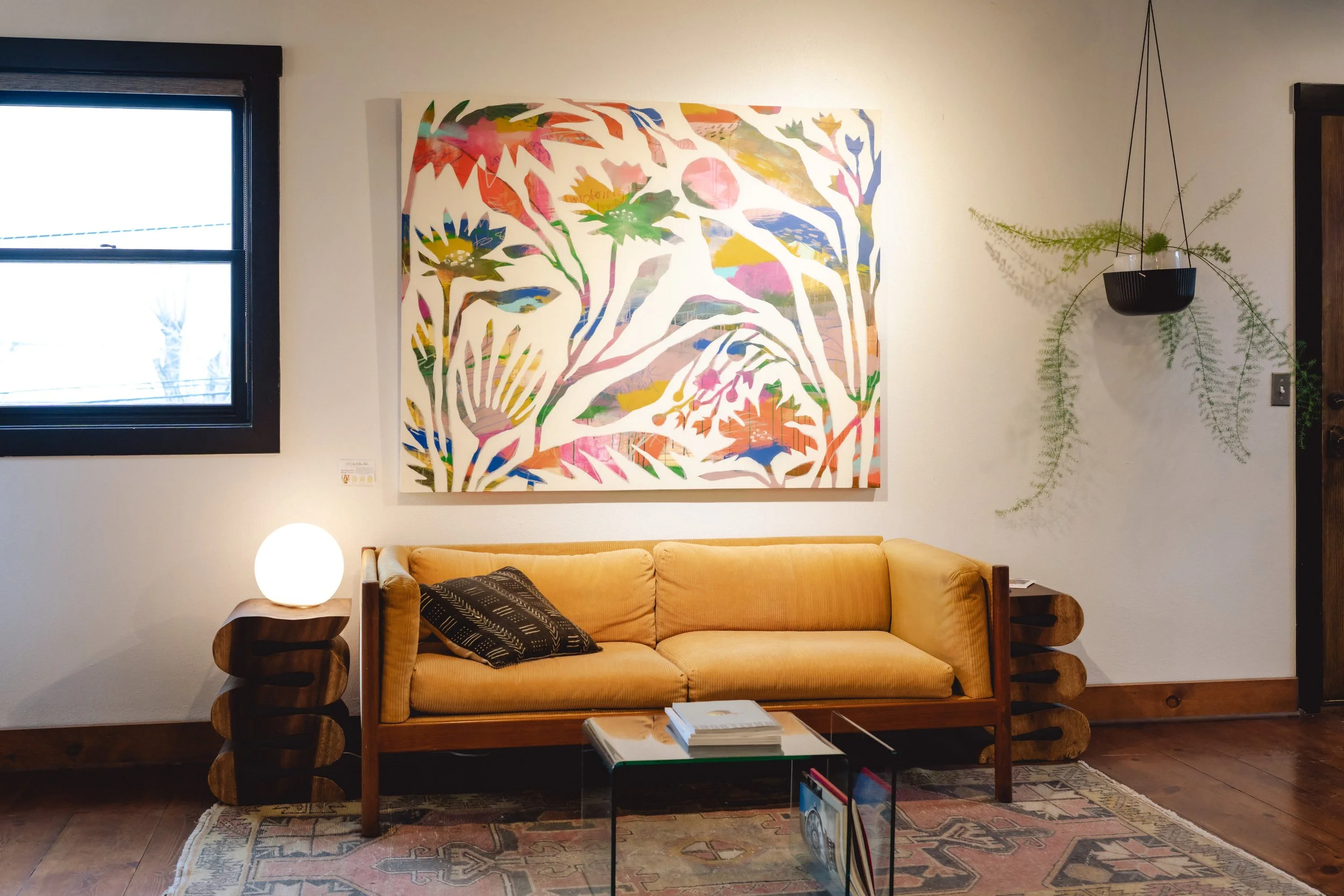 Living room with a yellow couch, a side table with a glowing lamp, abstract colorful wall art, a window with black trim, and a hanging plant on the wall.