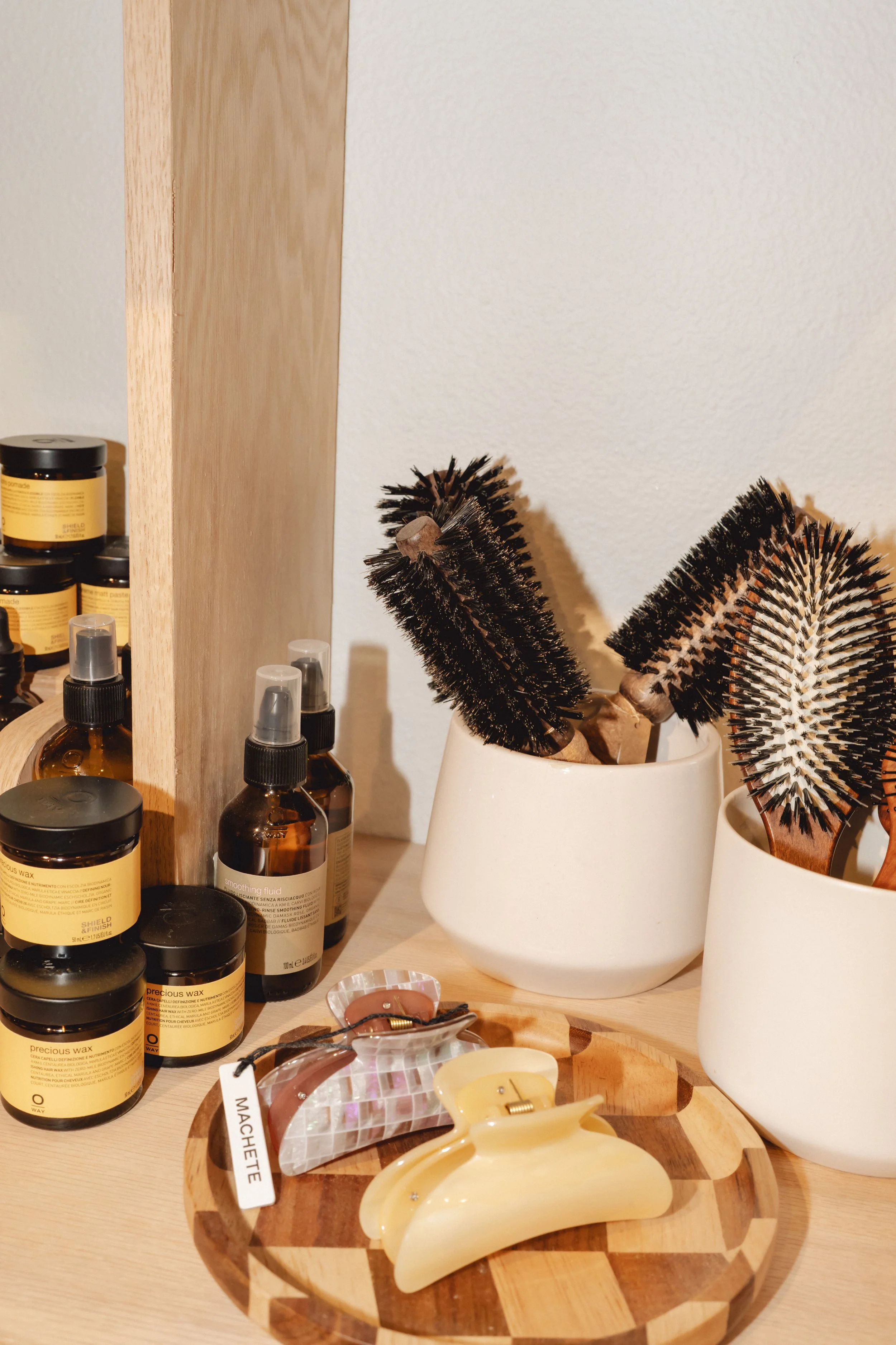 A wooden desk with hairbrushes in white ceramic holders, bottles of hair and skin products, and hair clips on a checkered wooden tray.