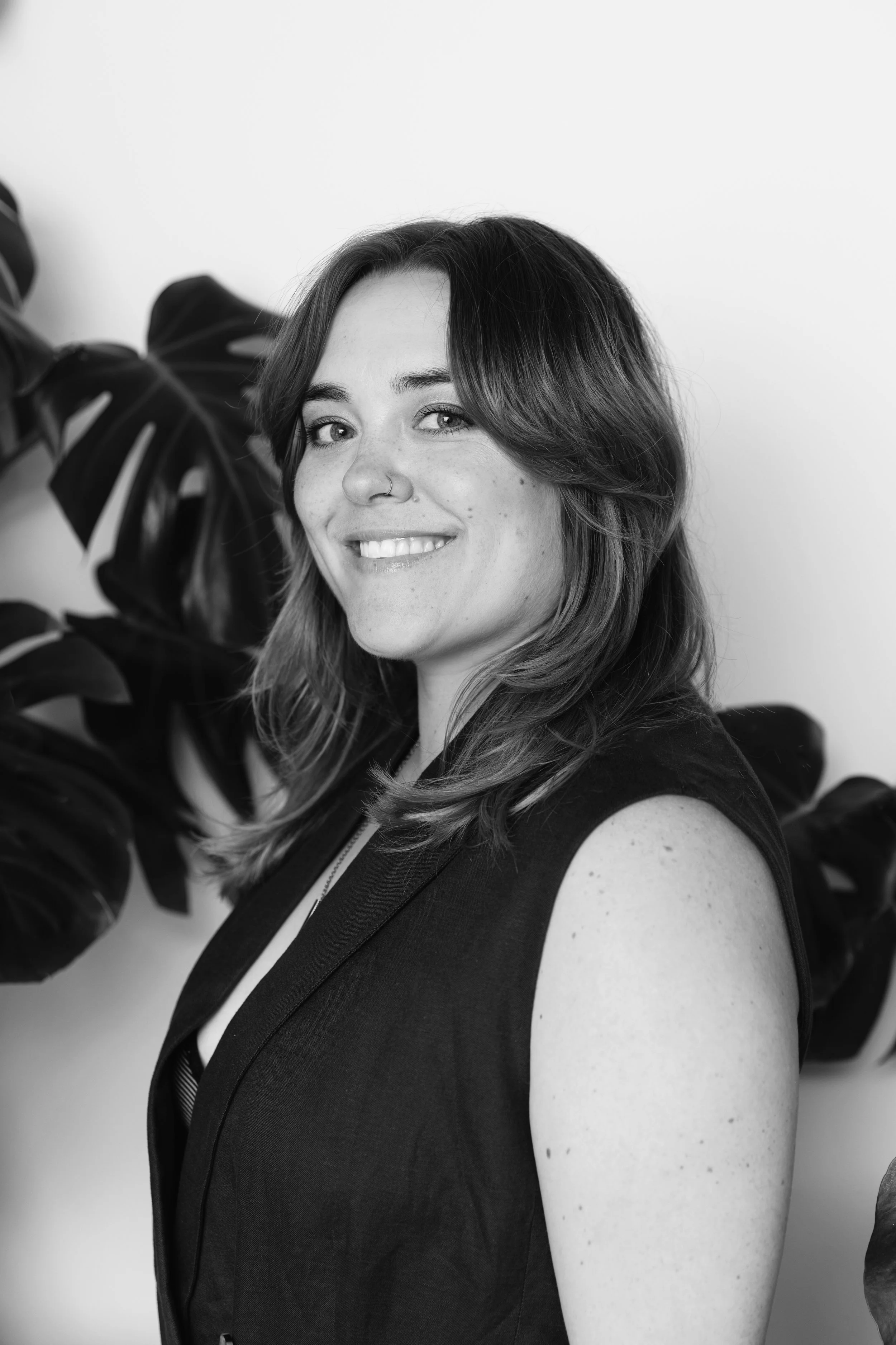 Black and white portrait of a woman smiling, with shoulder-length wavy hair, wearing a sleeveless dark top, standing in front of large dark leaves against a plain background.