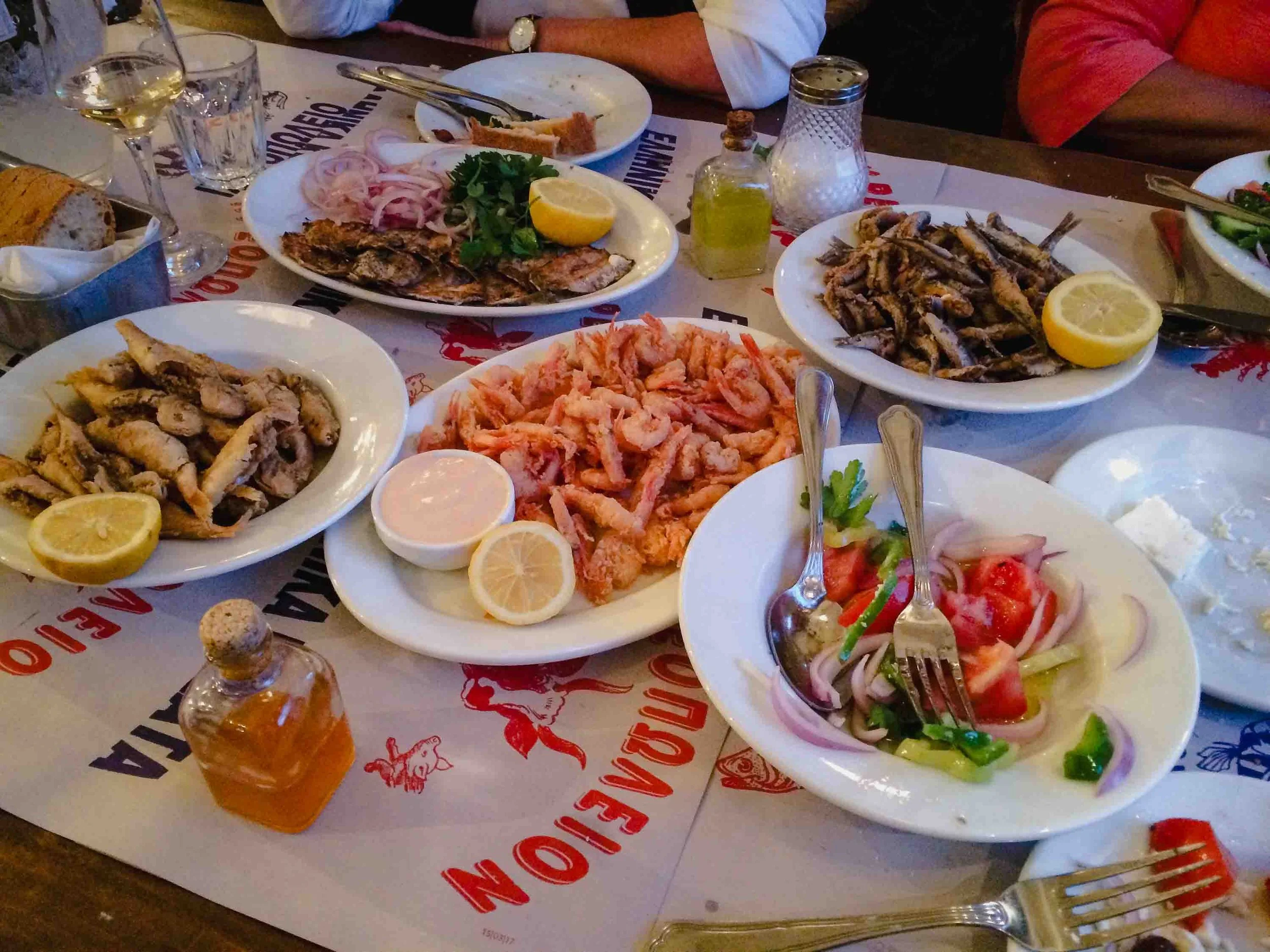 Traditional Greek mezze spread at an Athens taverna featuring fried calamari, grilled sardines, fresh shrimp, Greek salad with feta and Kalamata olives, and Greek wine on a branded table.