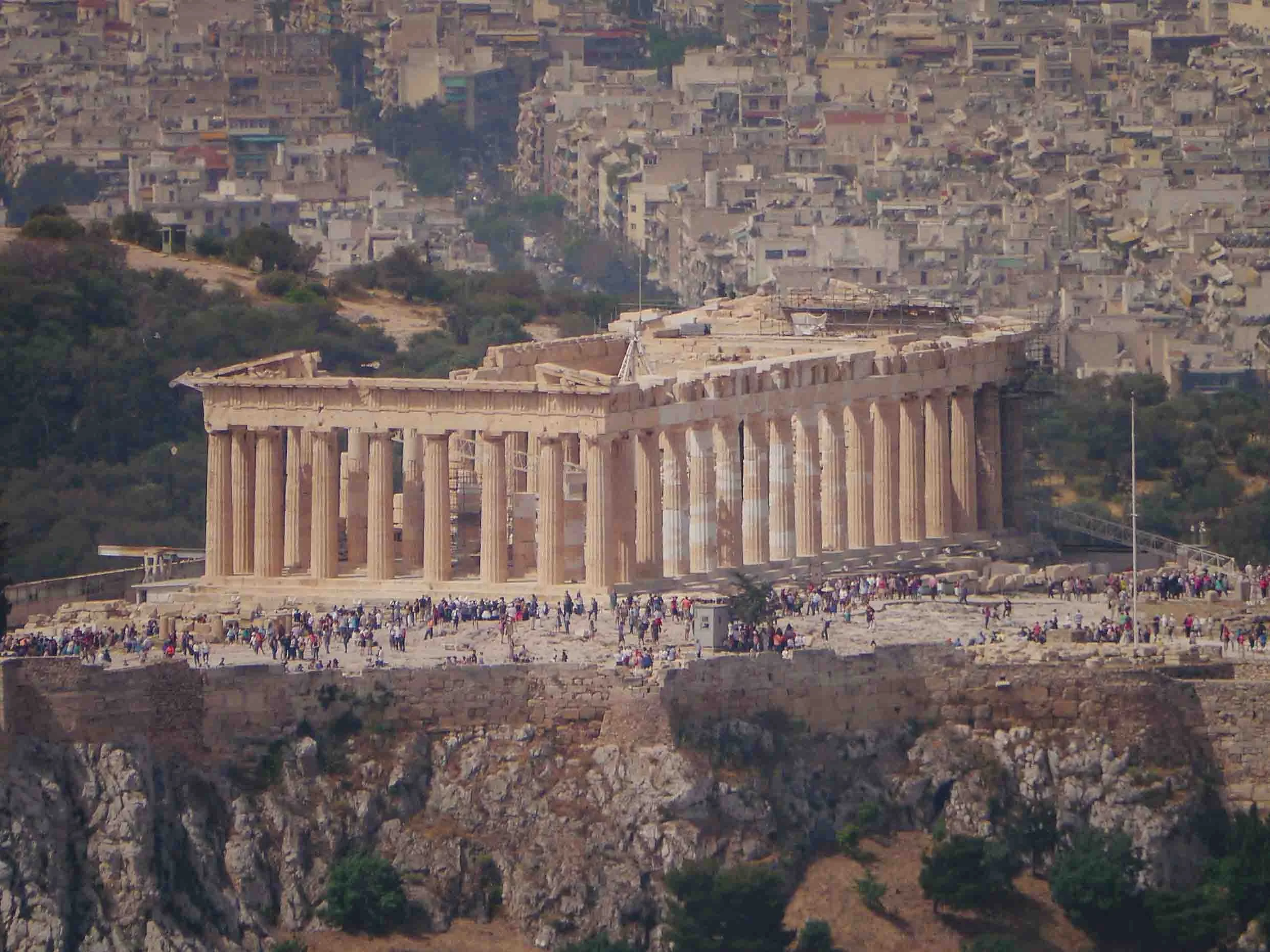 Panoramic view of the ancient Parthenon temple atop the Acropolis, surrounded by modern Athens cityscape, showing the iconic 5th-century BC structure with visitors exploring the archaeological site.
