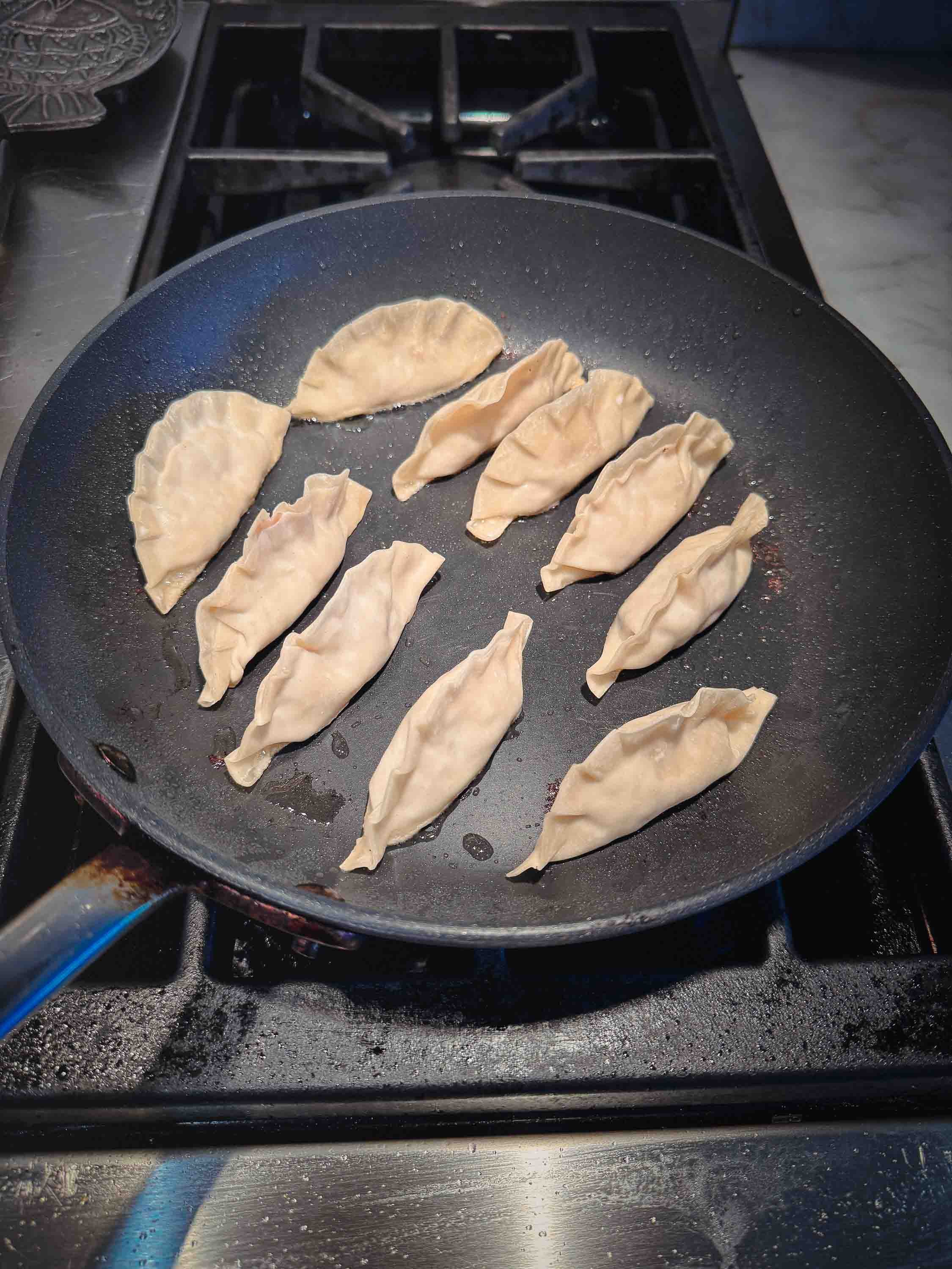 Homemade pork dumplings (pot stickers) cooking in a large non-stick skillet on a gas stove, showing the pleated wrappers before developing their signature crispy bottoms