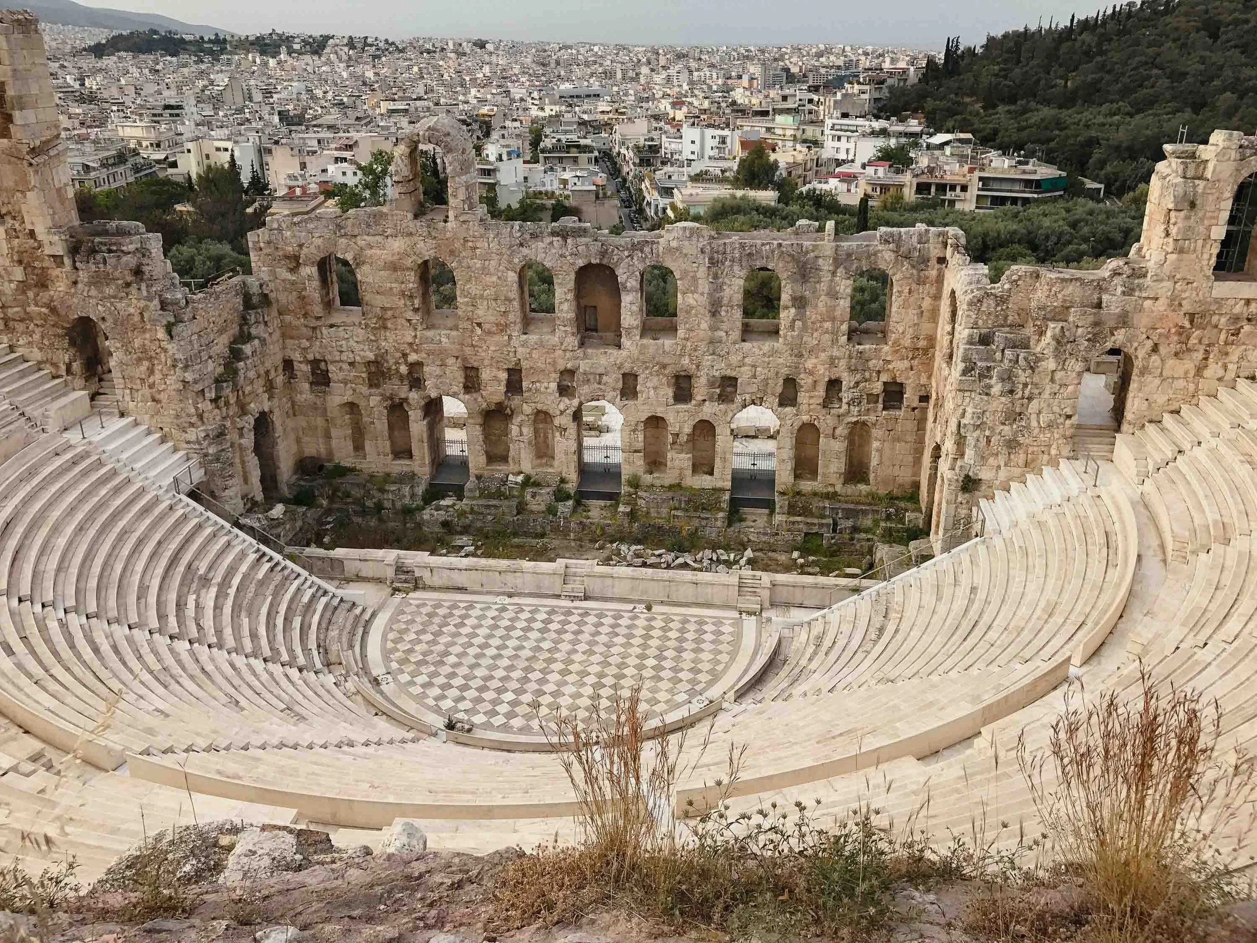 Ancient theater ruins at the Acropolis in Athens showing the semicircular stone seating tiers and weathered stone walls overlooking the modern city, demonstrating Greek architectural heritage.