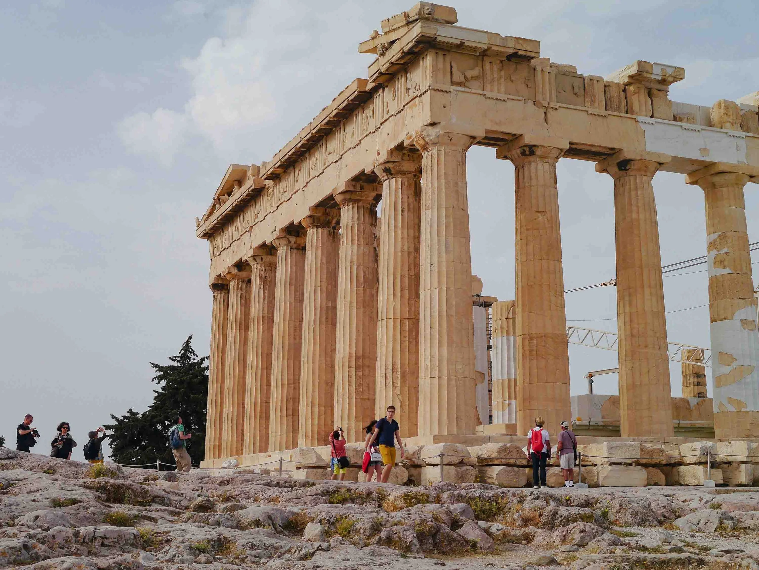 Close-up of the Parthenon's magnificent Doric columns on the Acropolis in Athens, with tourists exploring the ancient marble structure and restoration scaffolding visible on the temple roof.