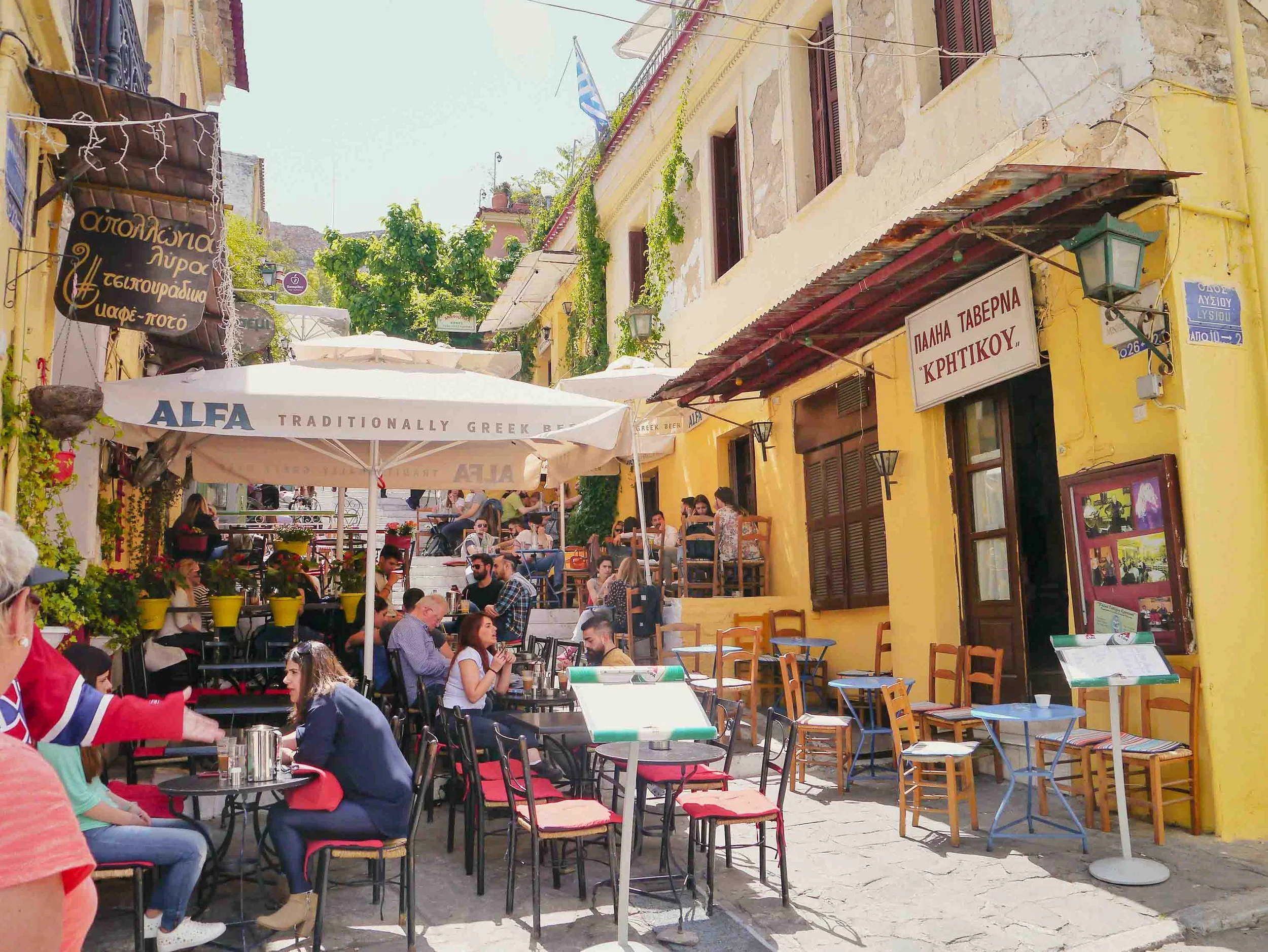 Bustling Athens taverna scene with outdoor seating on traditional steep steps, featuring white umbrellas and colorful tablecloths where diners enjoy Greek hospitality in a sunny neighborhood setting.