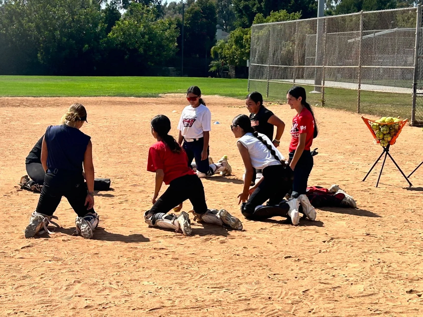 Group of young female softball players kneeling on a baseball diamond, receiving instructions from an adult coach during practice or a game. The players wear sports uniforms, and some have protective gear. There is a basket of yellow tennis balls in 