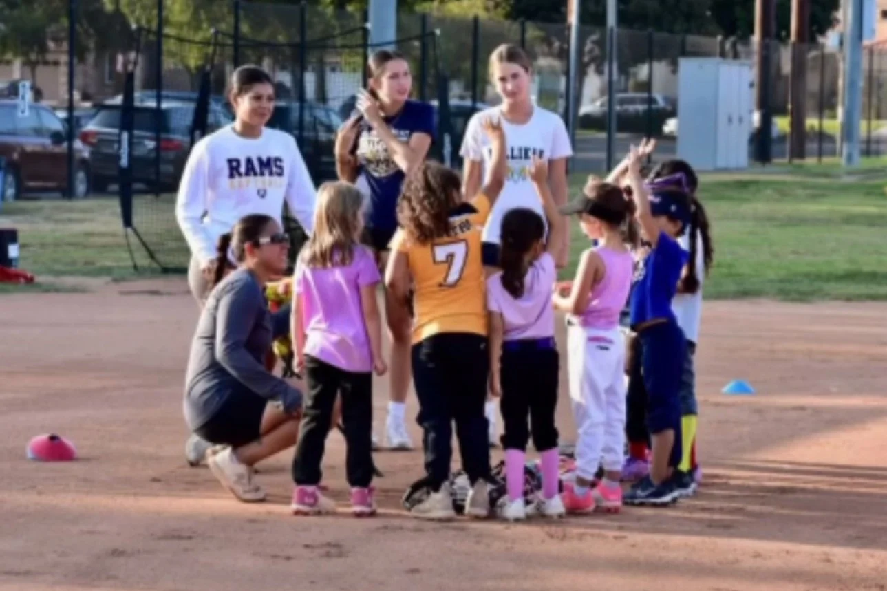 A group of young girls and women on a baseball field during a game, some wearing colorful uniforms, with one girl in a yellow jersey with the number 7. They are gathered in a huddle, engaging in a discussion or celebration, with some children raising