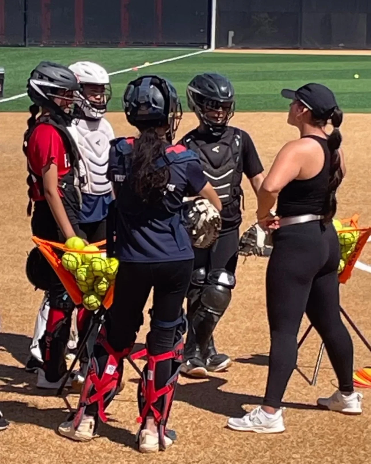 Group of female softball players and a coach having a discussion on the field during practice. The players are wearing helmets and catching gear, with some holding yellow softball balls. The coach is dressed in black athletic wear and wearing sunglas