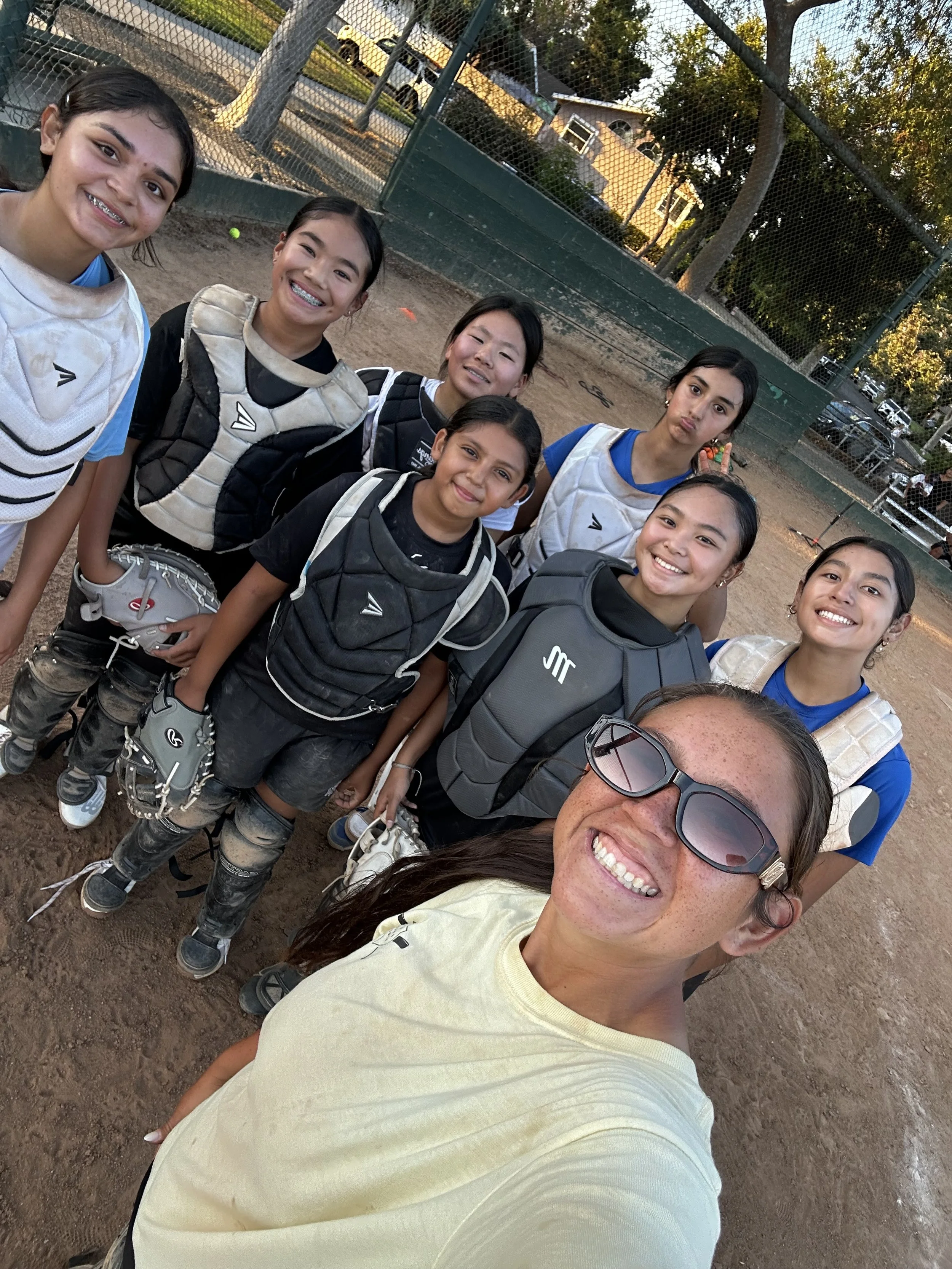 A group of women and girls smiling and posing for a selfie on a baseball field, with baseball gear including gloves and catcher’s gear. They are dressed in sports uniforms, and some are making peace signs or playful faces.
