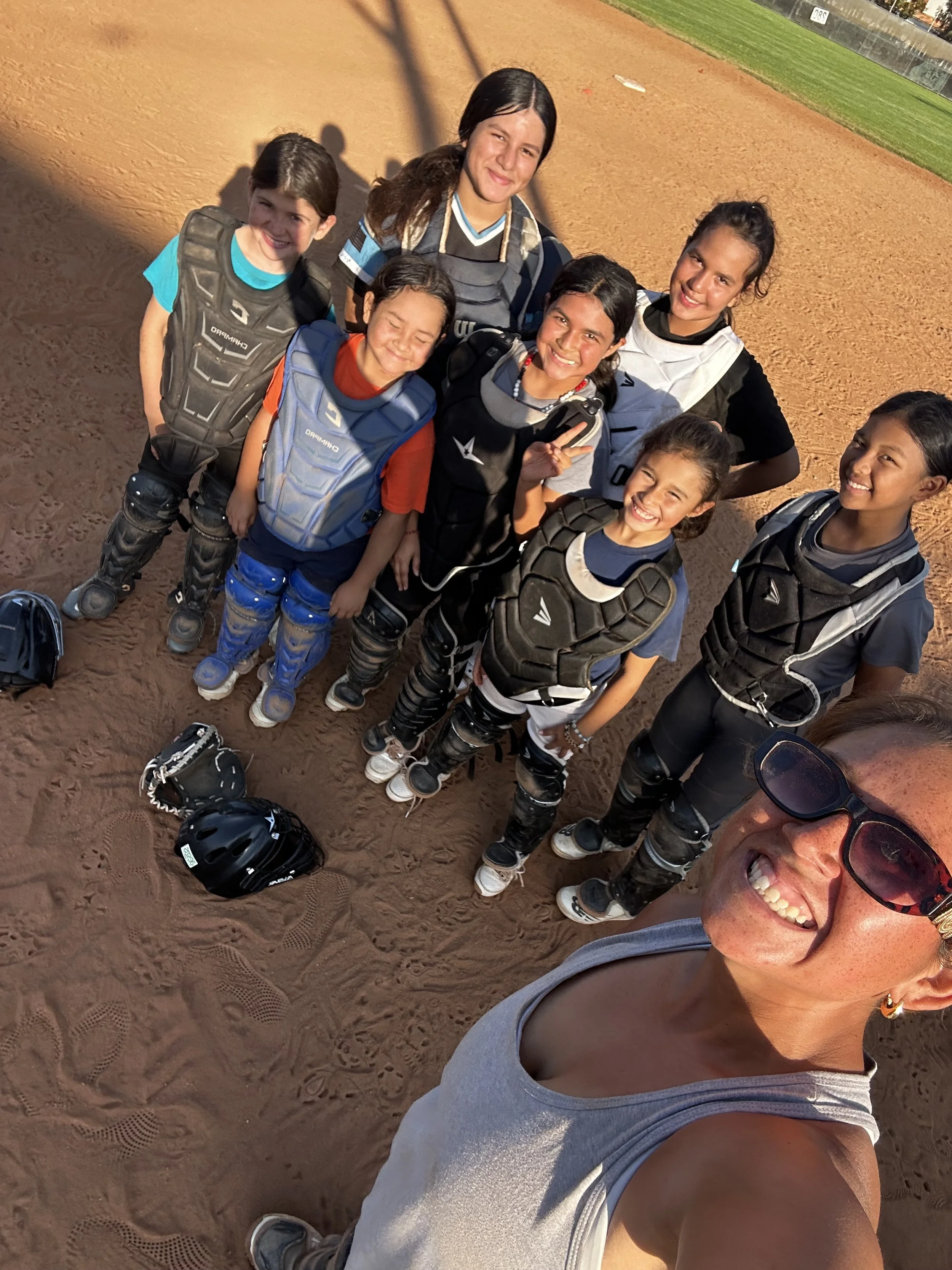 A group of children and a woman taking a selfie on a baseball field in the late afternoon. The children are dressed in baseball uniforms and catcher's gear, and they are smiling.