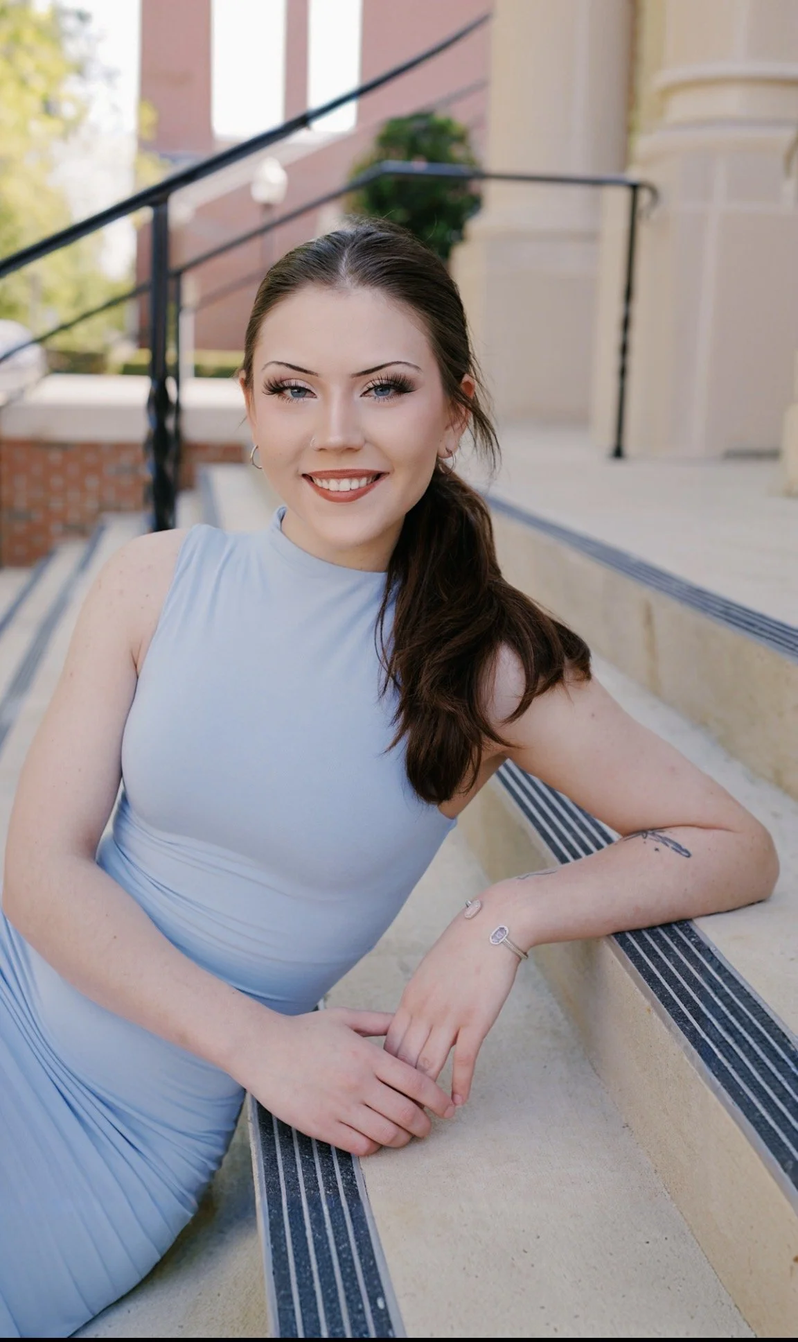 A woman with long dark hair, blue eyes, and makeup, smiling, wearing a sleeveless light blue dress, leaning on a railing outside of a building with stairs and brick and beige walls.