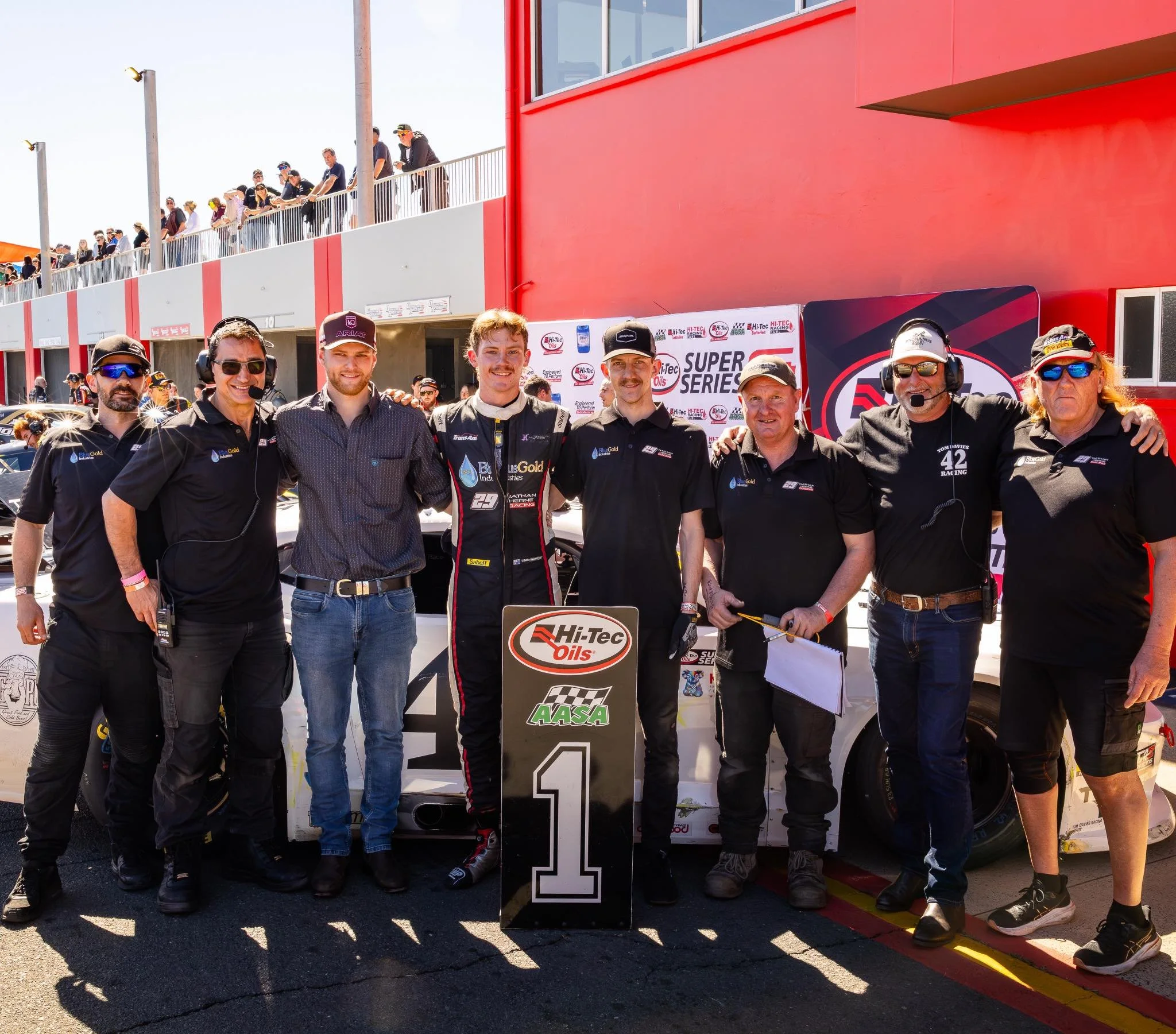 Race car driver and team members celebrating on a racing track with a winner's sign reading '1' in front of a race car, with spectators and a grandstand in the background.