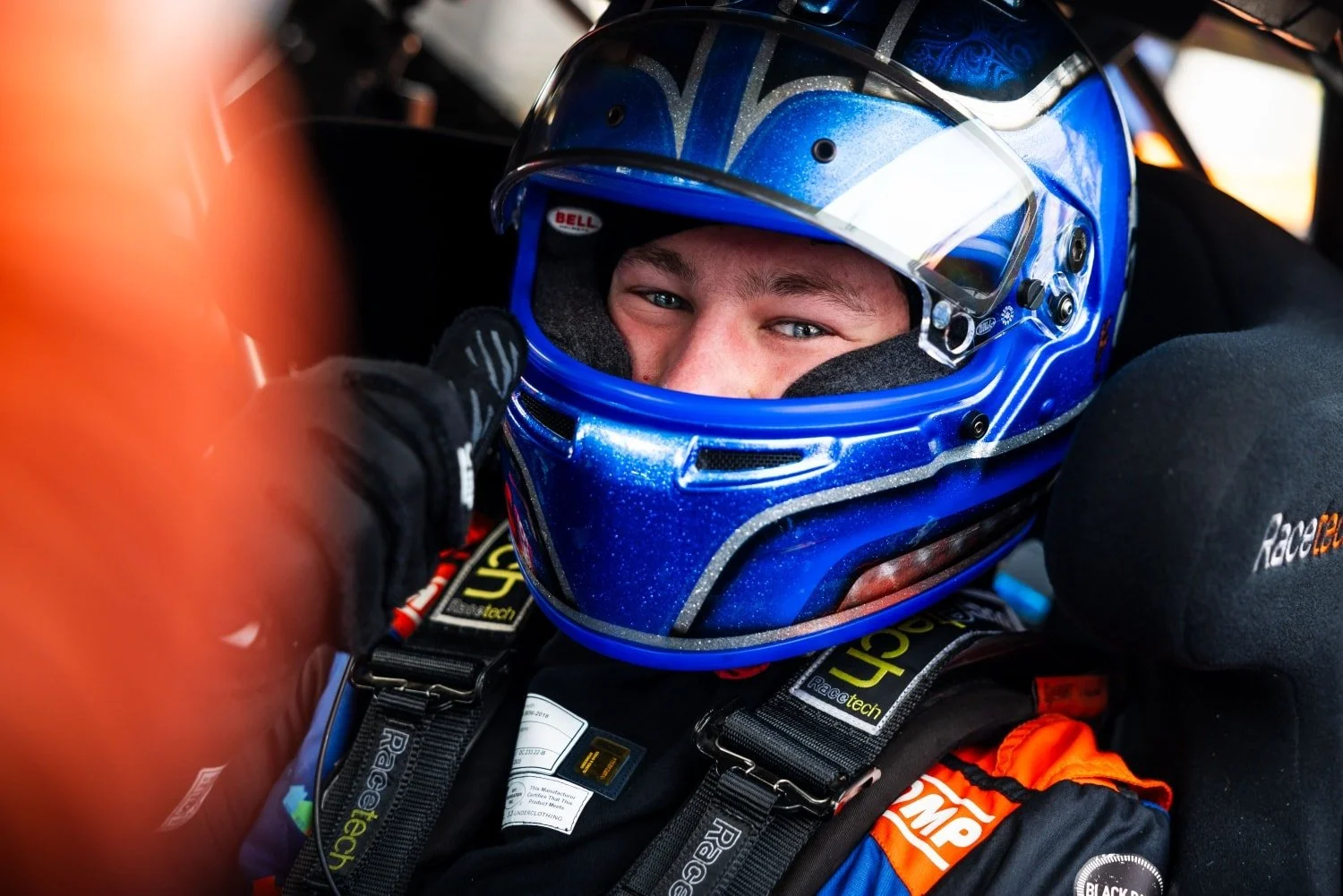 Close-up of a race car driver wearing a blue helmet, sitting in the cockpit, and making a gesture with their hand.