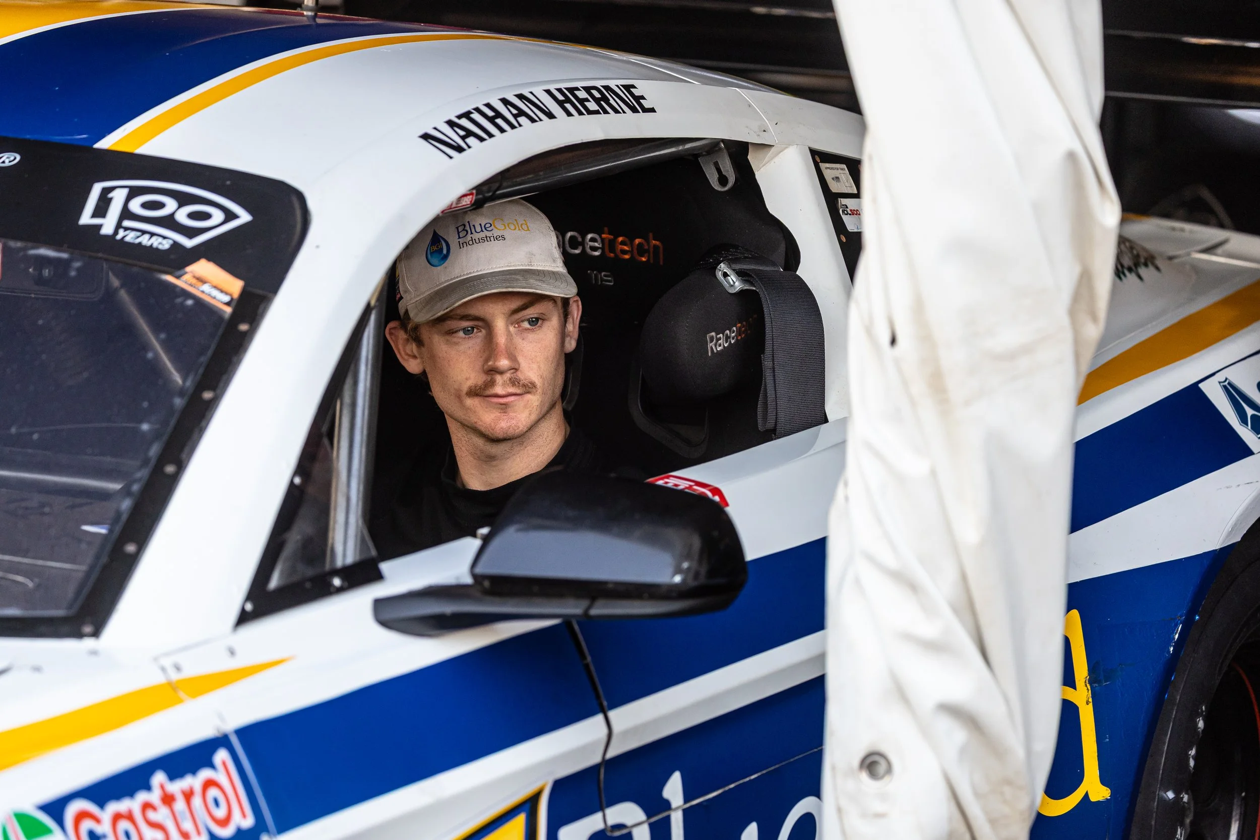 Race car driver sitting inside a blue and white race car with the name Nathan Herne on the windshield, wearing a cap and looking to the side.