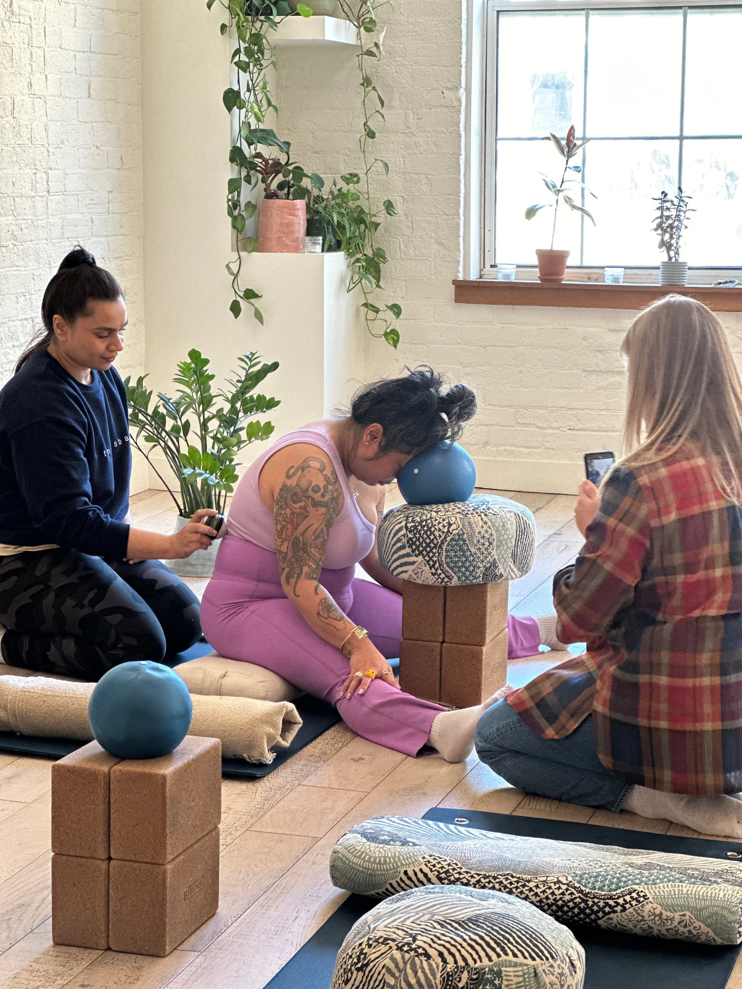 Three people practicing yoga in a bright room with plants. One person is in a seated position, resting their head on a yoga block and cushion, while others assist and observe. Yoga props are arranged around them.