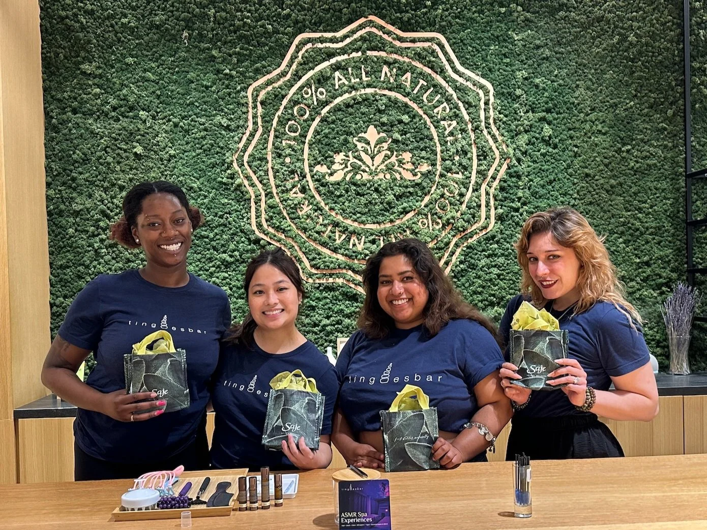 ASMRtists smiling and holding gift bags in front of a green moss wall with "100% All Natural" text, in a store setting with aromatherapy products on the counter.