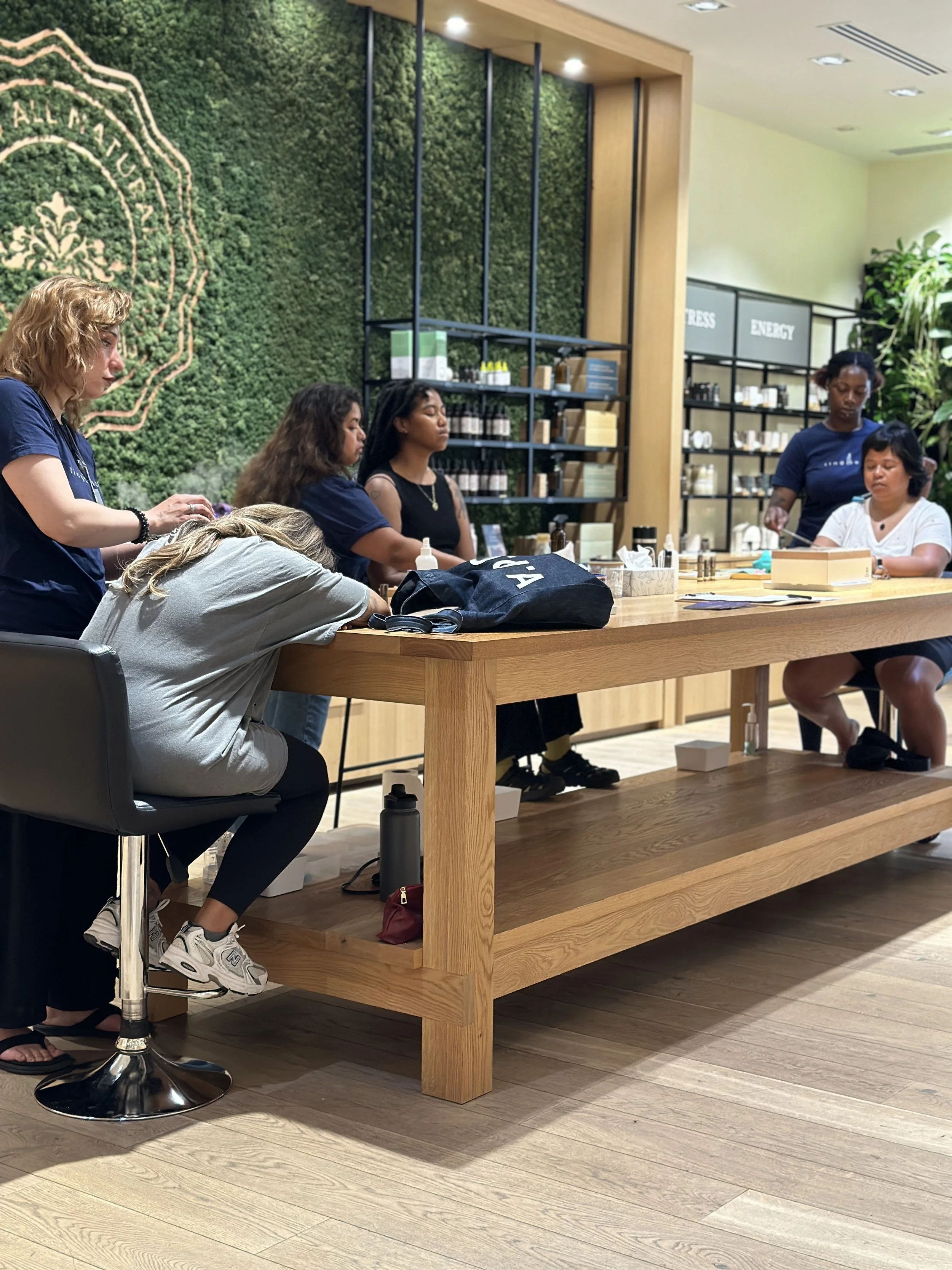 Group of women sitting around a wooden table, some are resting on it, located inside a store with shelves and a green wall backdrop.
