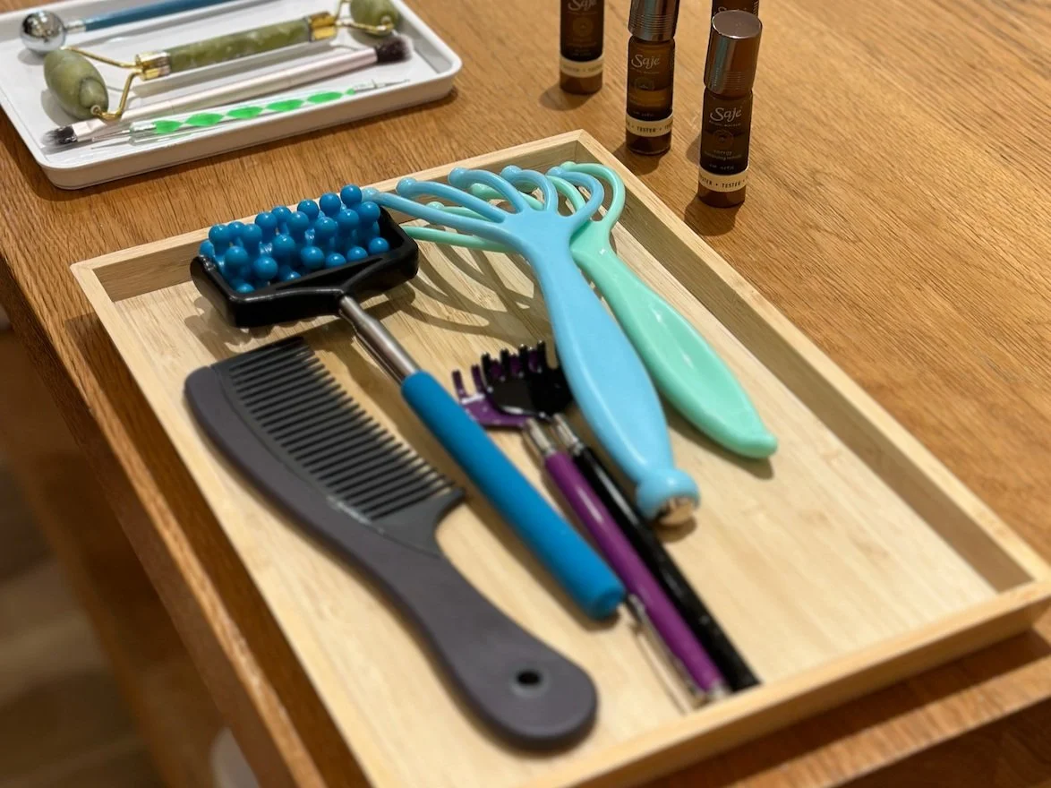 A wooden tray on a table containing various massage tools, a black comb, and hairbrushes. Nearby, there are bottles of essential oils and a tray with jade rollers and brushes.
