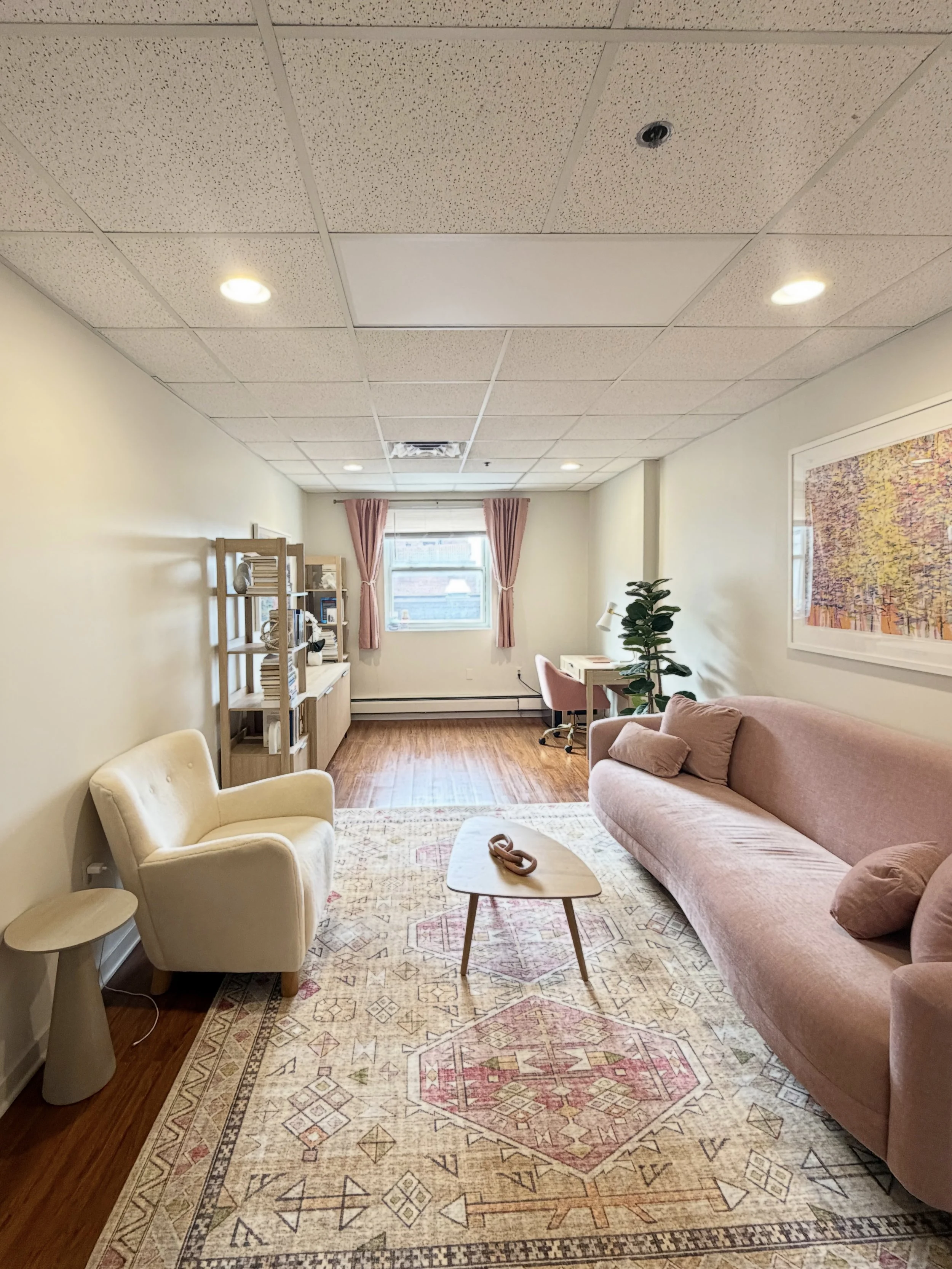 Living room with a pink couch, cream armchair, wooden coffee table, patterned rug, bookshelf, window with pink curtains, painting on the wall, and potted plant.