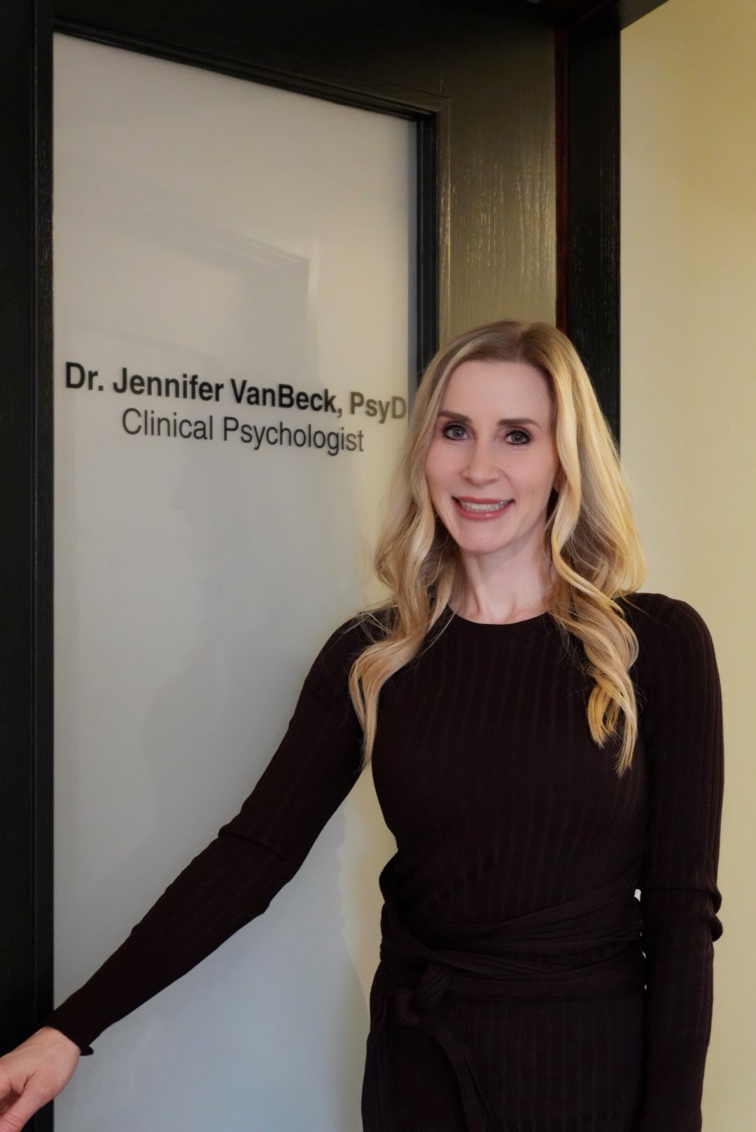 A woman with blonde hair and a black long-sleeve dress standing next to a door with a sign that reads Dr. Jennifer VanBeck, PsyD, Clinical Psychologist.