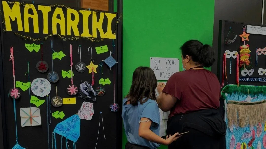 A girl and a woman are standing at a sign that reads 'Put Your Art Up Here' at an art display area decorated with handmade crafts, including paper ornaments, stars, and masks.