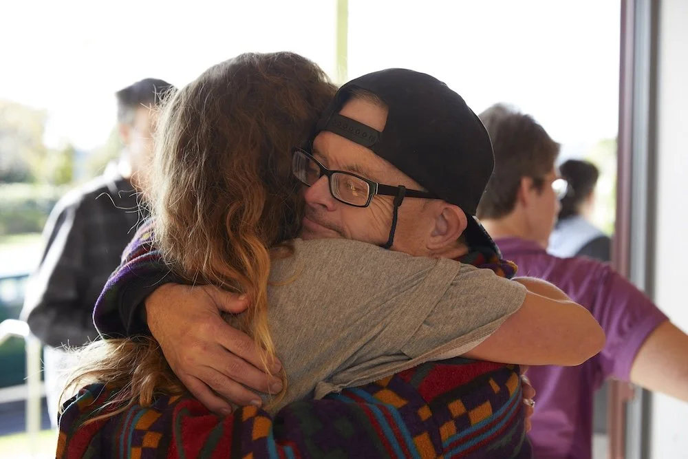 A man and woman embracing in a hug indoors, with other people blurred in the background.