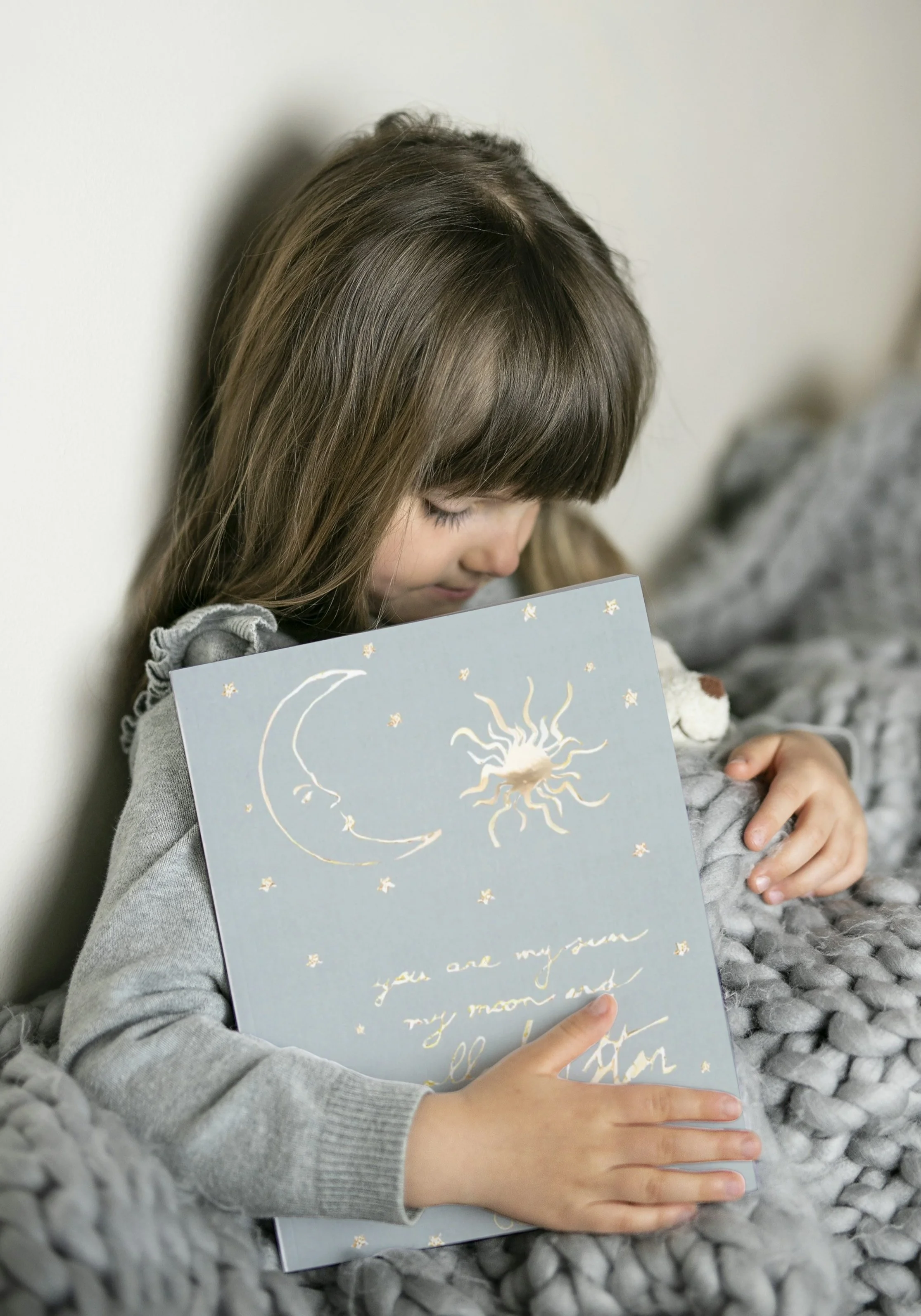 A young girl sitting against a wall, holding a decorative card with moon and star illustrations and gold writing, and hugging a plush toy.