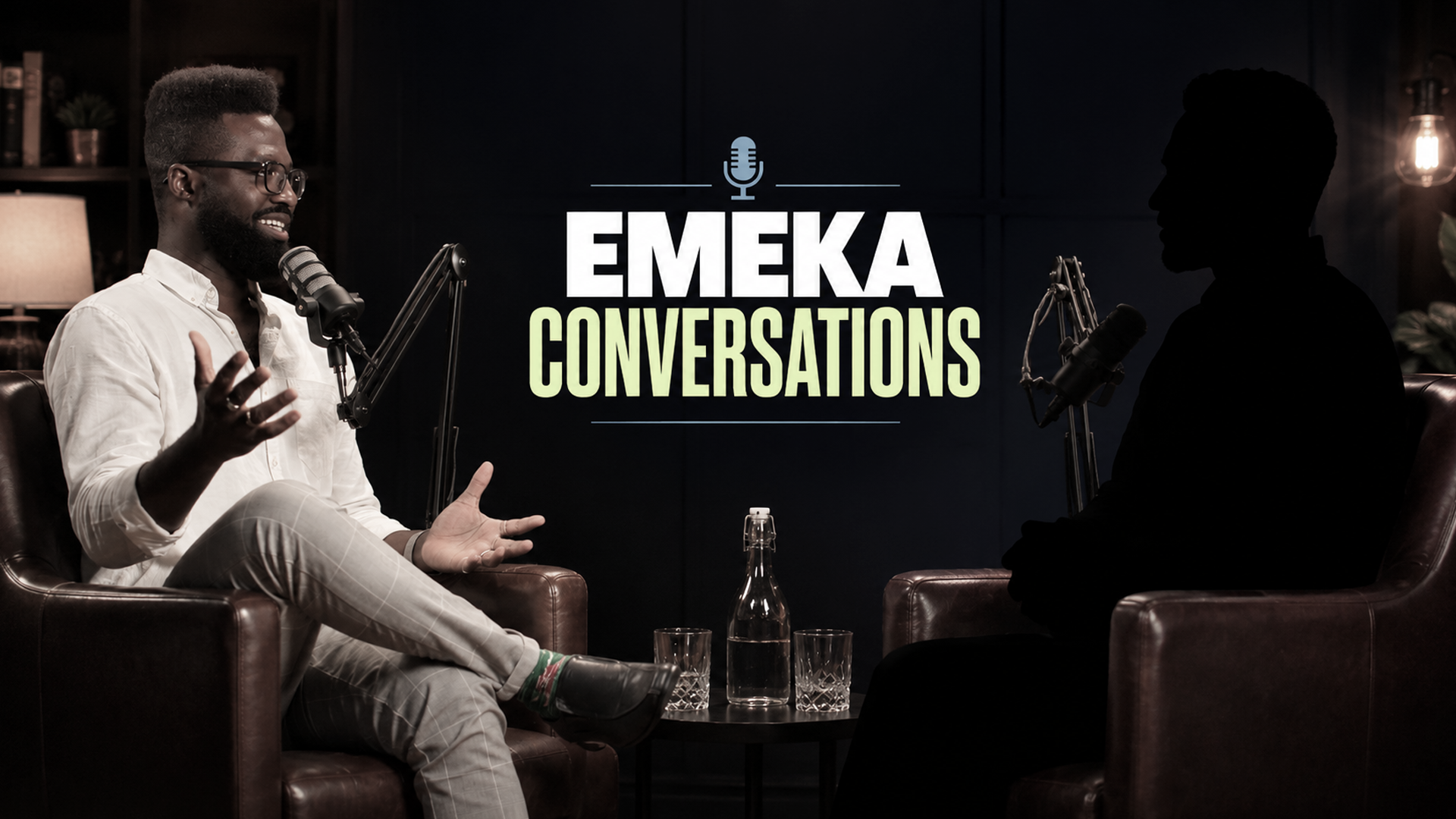 Two people having a conversation in a recording studio with microphones. Emeka is wearing a white shirt and glasses, smiling and gesturing with his hands. The other person is a silhouette. A sign reads "Emeka Conversations" with a microphone icon.