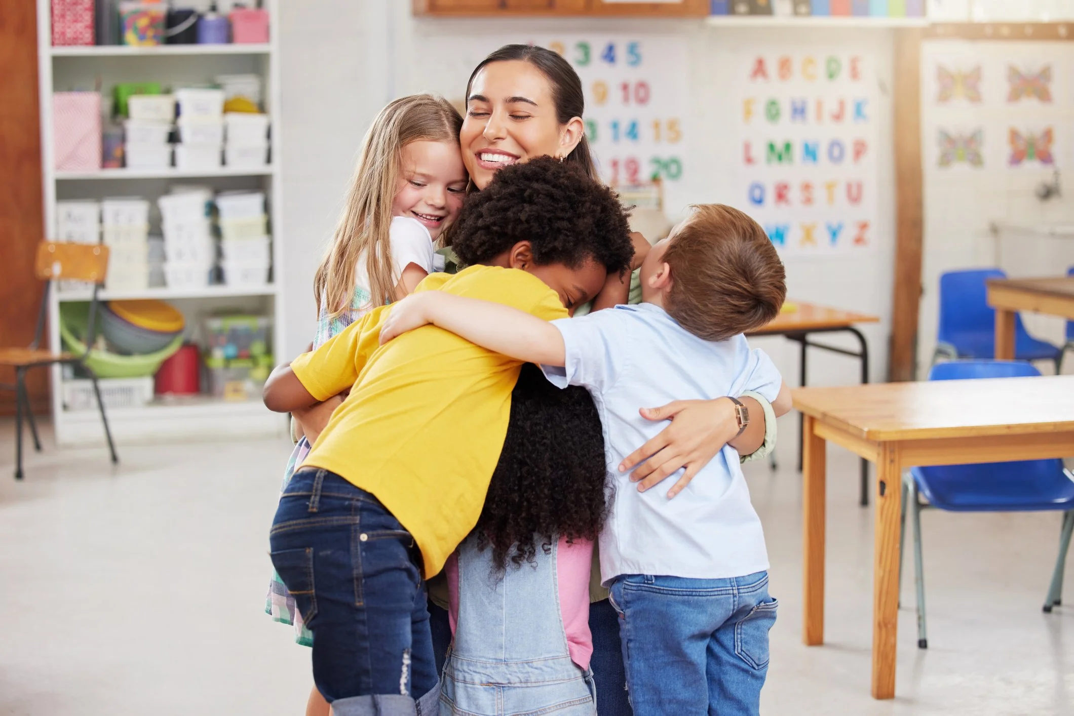 A group of children and a woman hugging and smiling in a classroom with educational posters and shelves in the background.