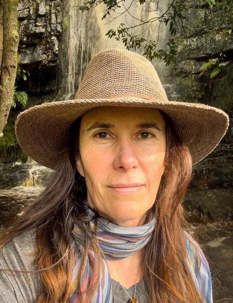 A woman wearing a wide-brimmed straw hat and a multicolored scarf around her neck, standing outdoors near a waterfall and lush greenery.
