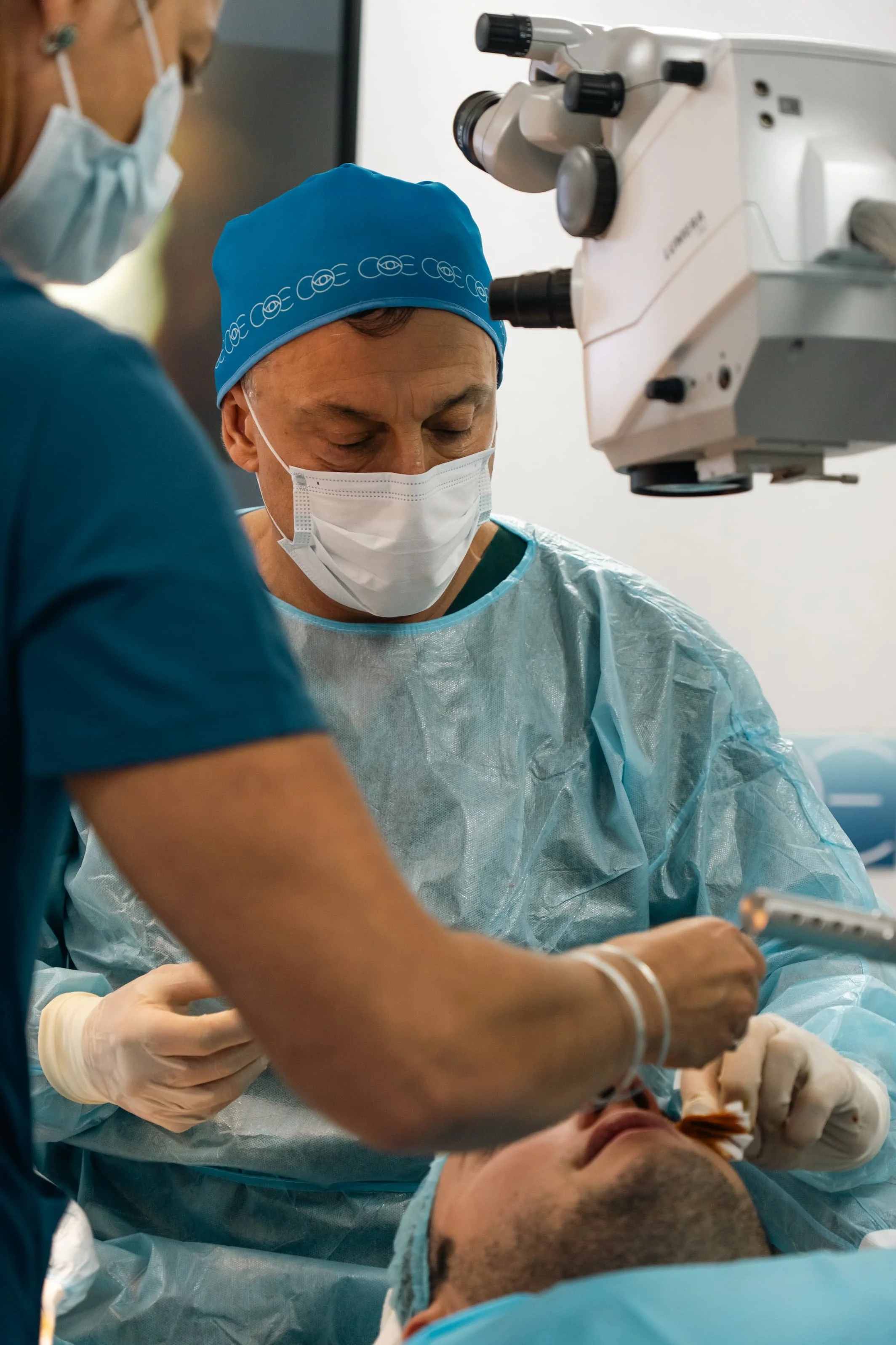 Médicos en quirófano realizando una cirugía con ayuda de un microscopio, ambos con mascarillas y batas quirúrgicas.