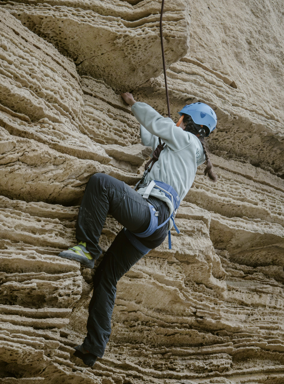 Person climbing a rock wall, wearing a blue helmet, climbing harness, and outdoor clothing, on a textured sandy-colored rock formation.
