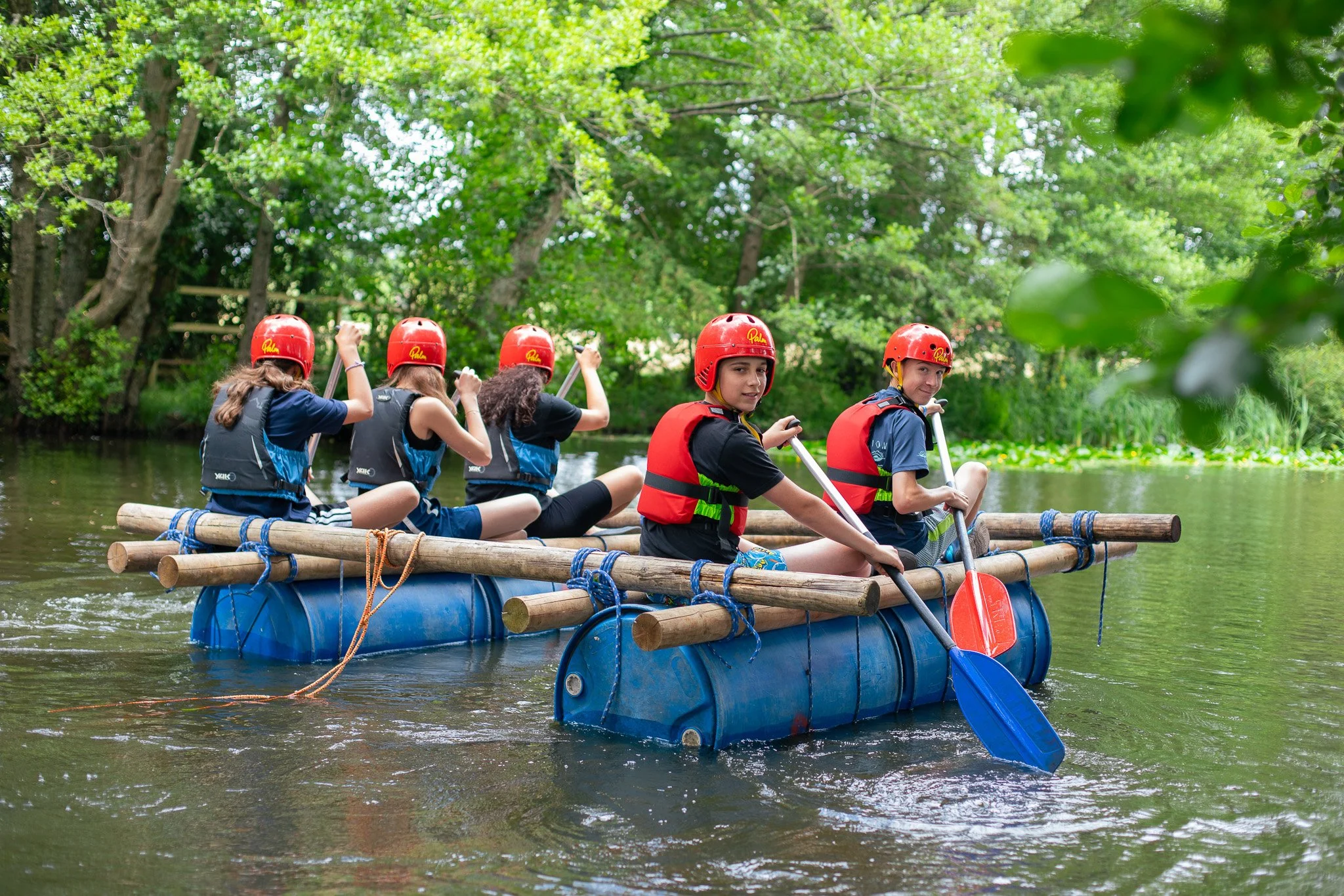 Children wearing life jackets building a raft with barrels, ropes, and wooden planks by a lake during a summer camp activity.