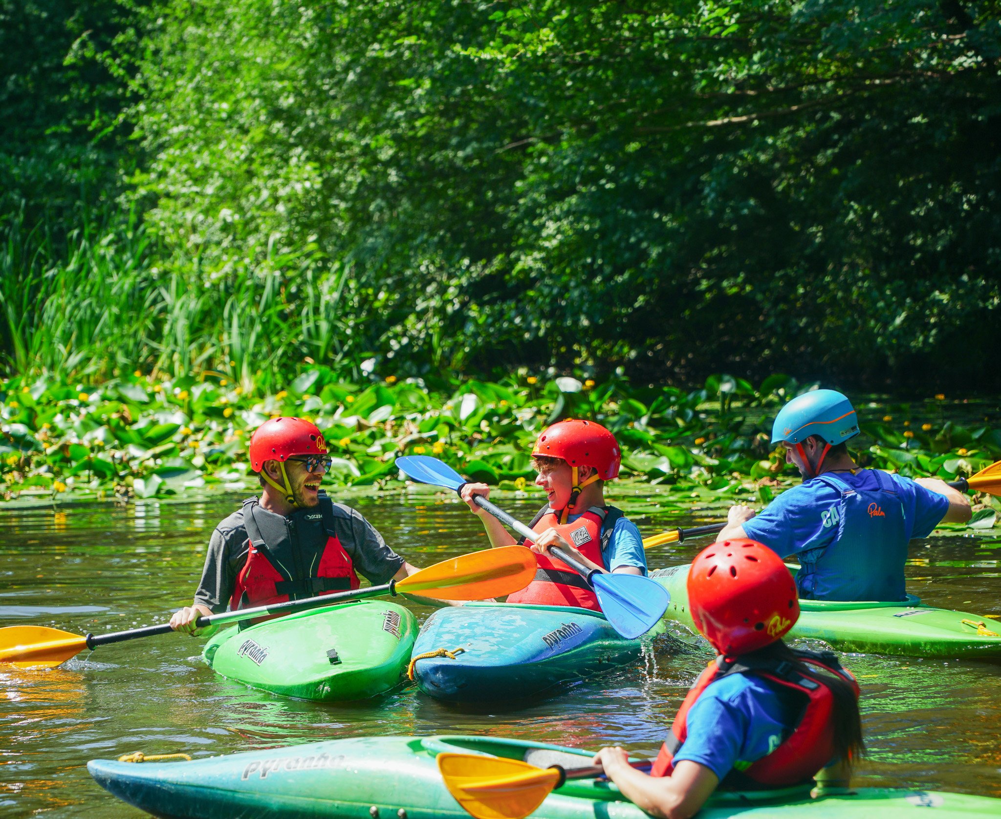 A child wearing a helmet and life jacket paddling a kayak on a calm lake during a summer camp activity.