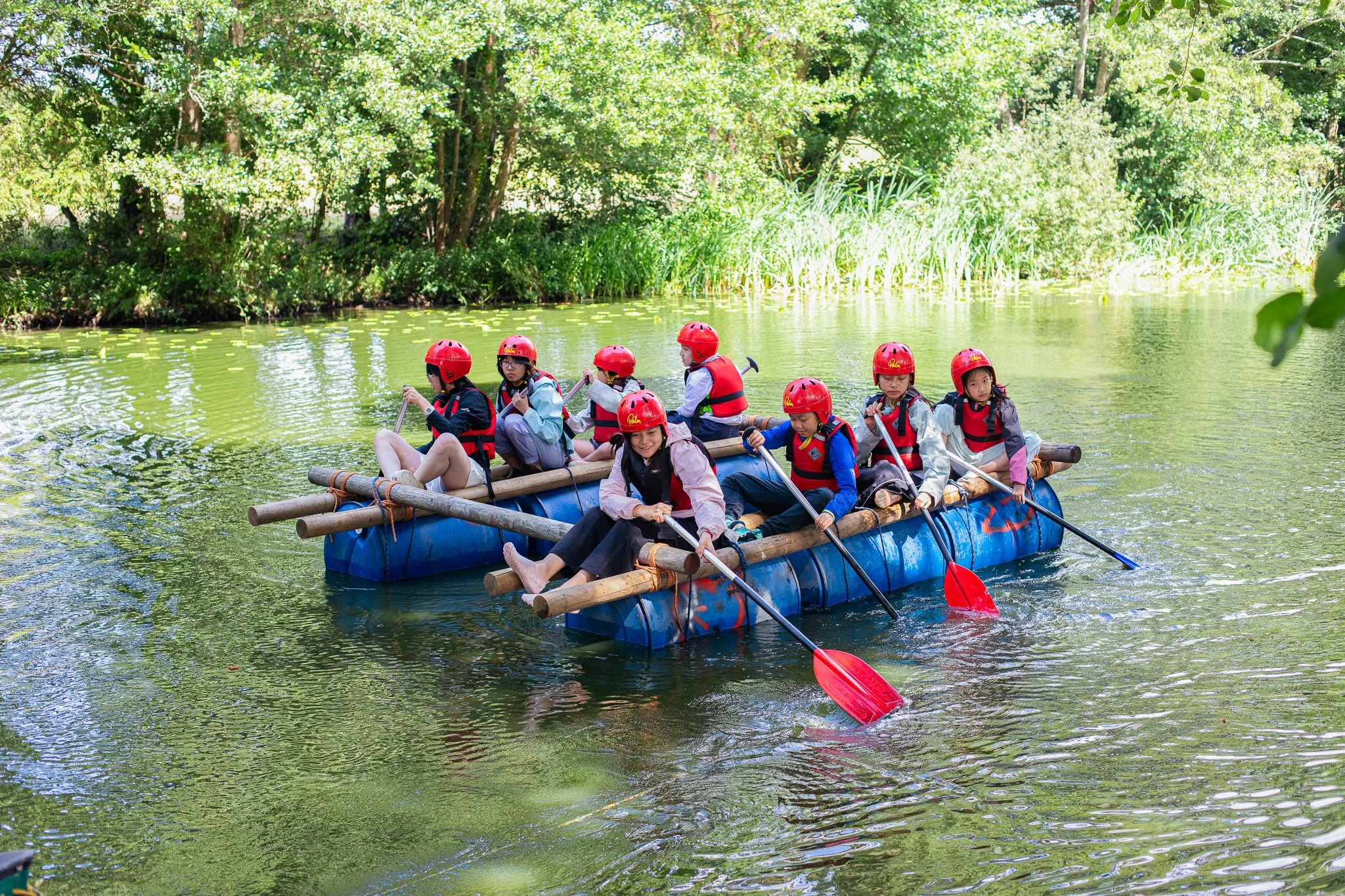 A group of teens in front of a climbing wall outdoors, all wearing safety helmets and smiling for the photo.