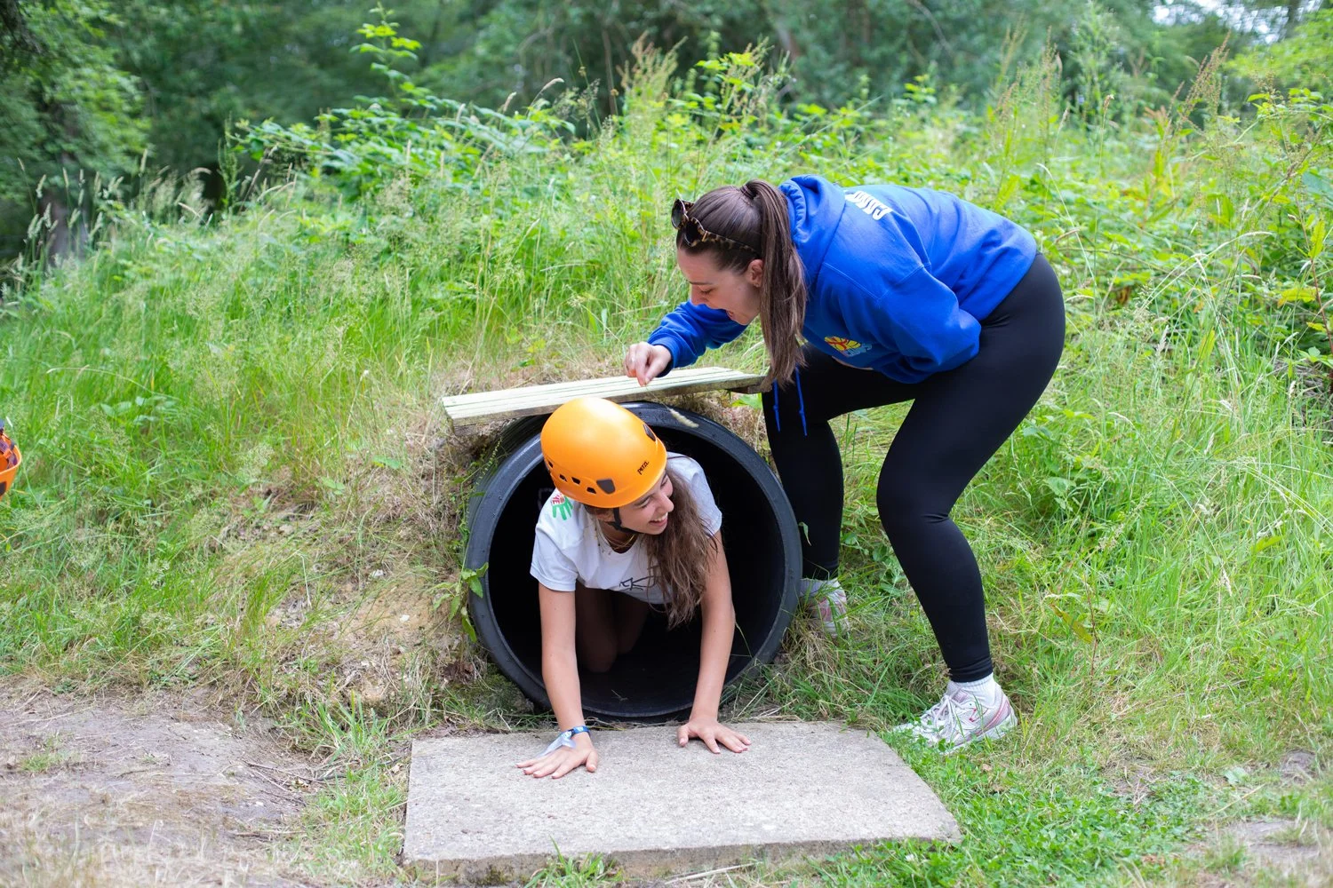Child wearing a helmet and safety gear crawling through an outdoor adventure camp tunnel system.