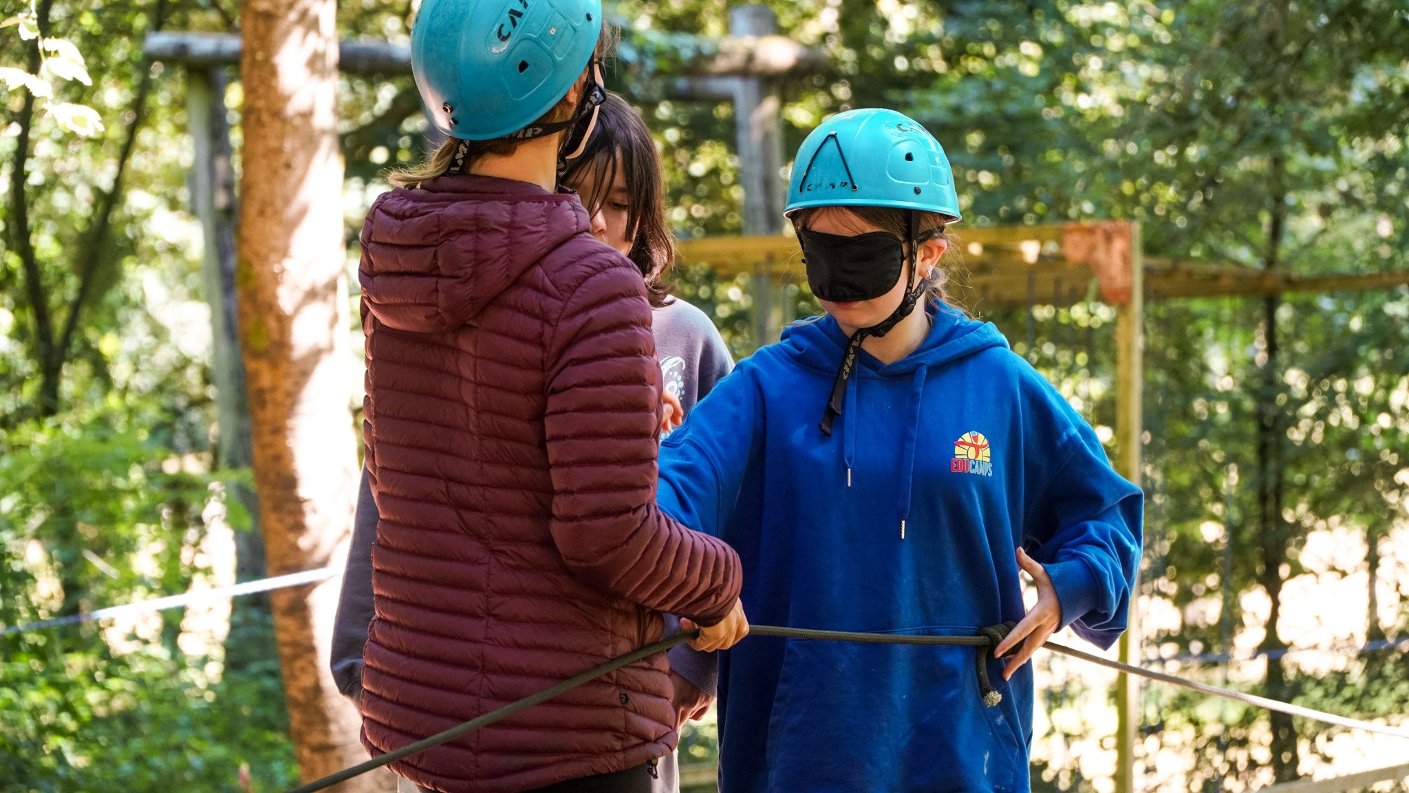 Children wearing blindfolds, holding onto a rope, and navigating an outdoor Nightline obstacle course together.