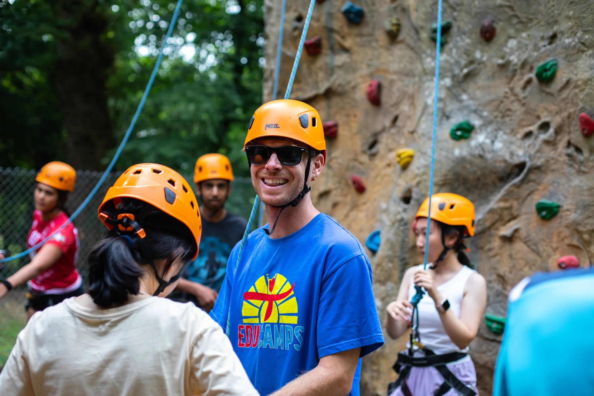 Group of people wearing orange helmets and harnesses at an outdoor rock climbing wall in a forested area. The instructor is teaching a climbing wall activity on a residential adventure camp.