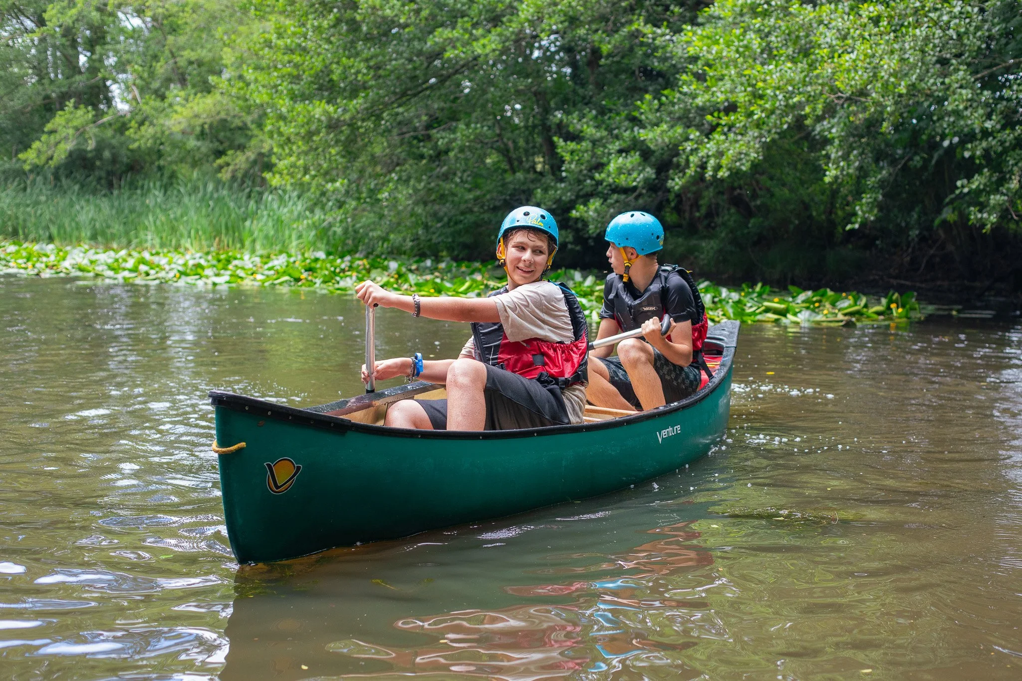 Group of people wearing life jackets in green canoes on a lake, surrounded by trees and other boats, under a partly cloudy sky.