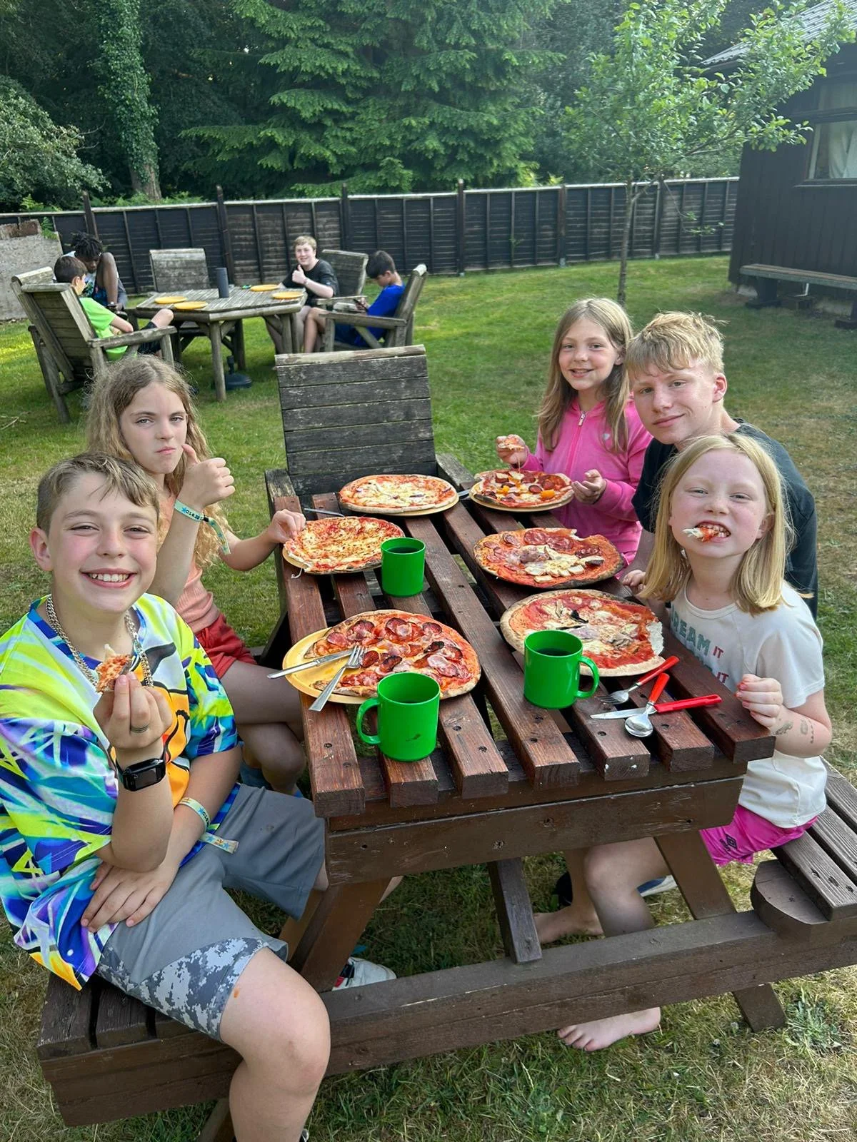 A group of children sitting at an outdoor picnic table with large pizzas and green cups. They seem to be enjoying a meal in a backyard or garden setting with trees and a wooden fence in the background. Some children are smiling and holding slices of pizza.