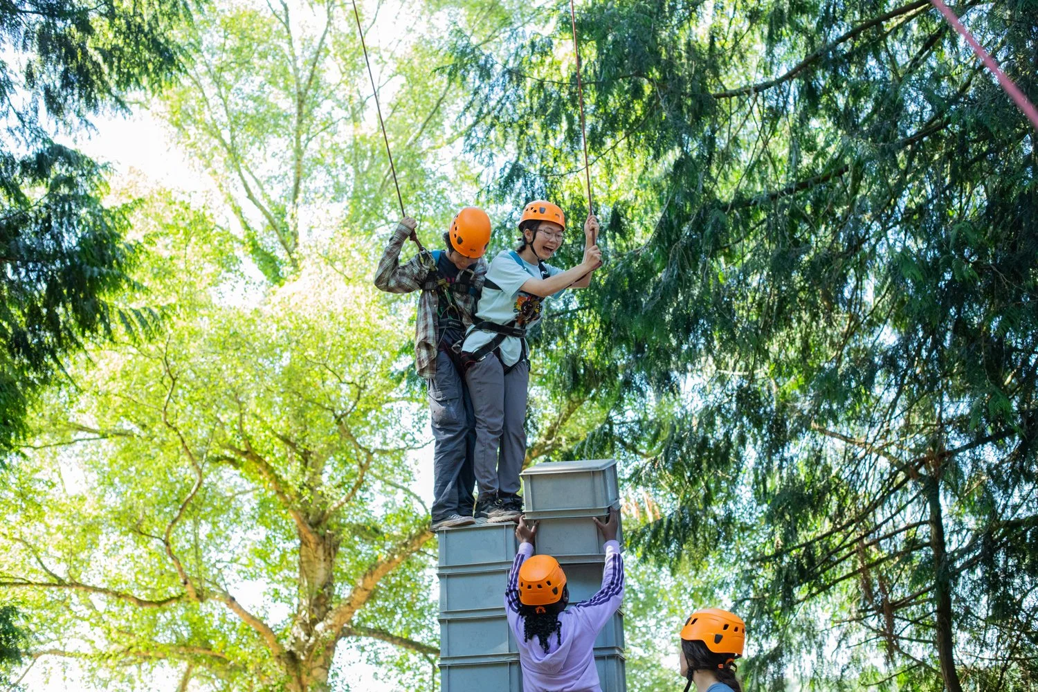 Two children wearing helmets and harnesses balancing on a tall stack of crates, supported by ropes, during a team-building activity at summer camp.