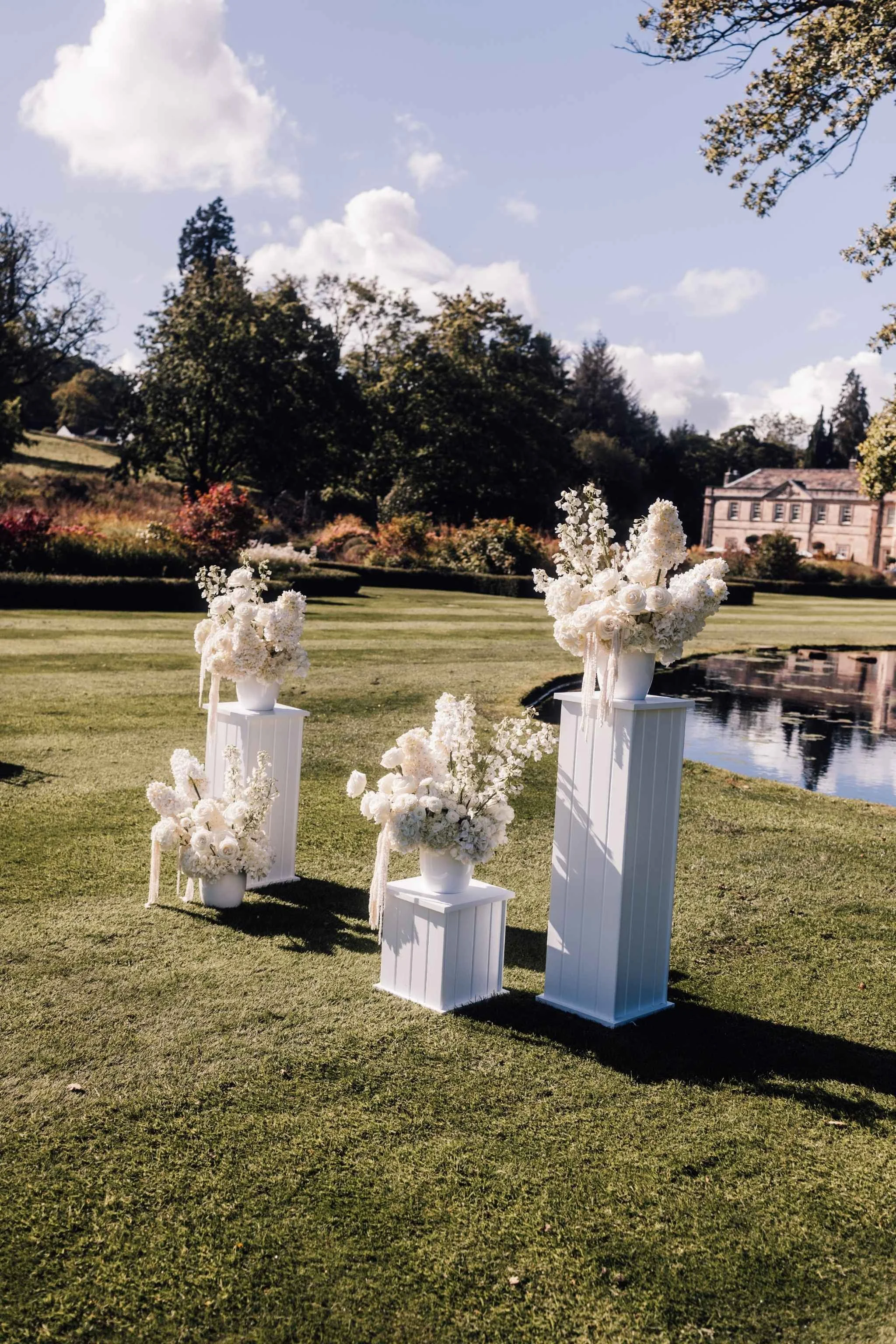 Outdoor wedding altar decorated with white flowers and floral arrangements on white pedestals, set on a grassy lawn with a pond and trees in the background.