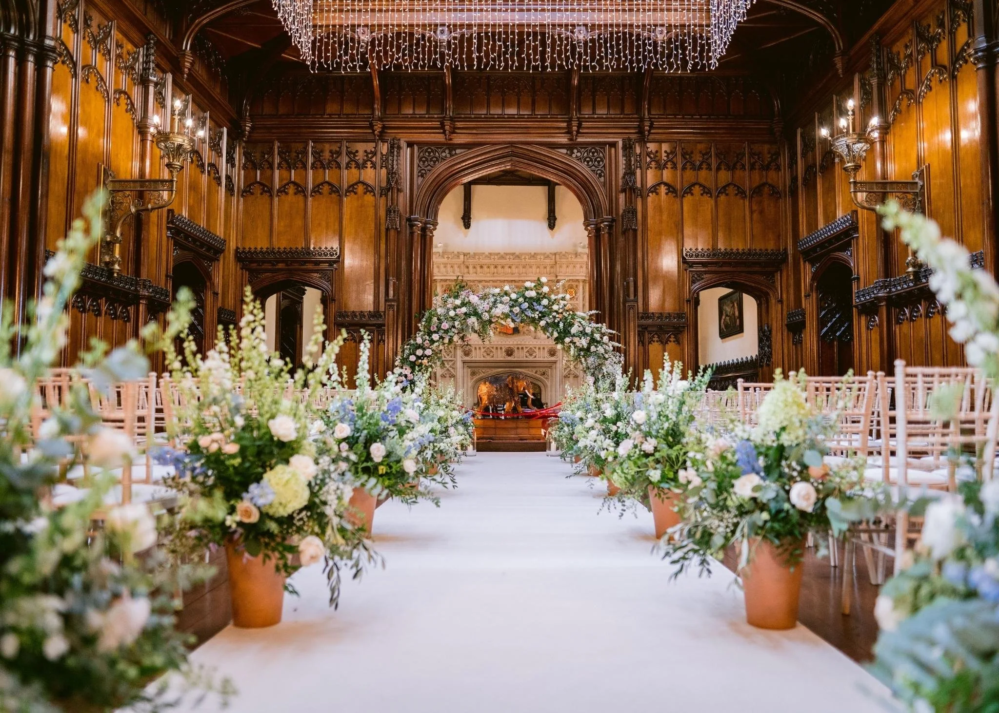 A decorated wedding aisle in an ornate wood-paneled hall with a floral arch at the altar and rows of chairs on either side.