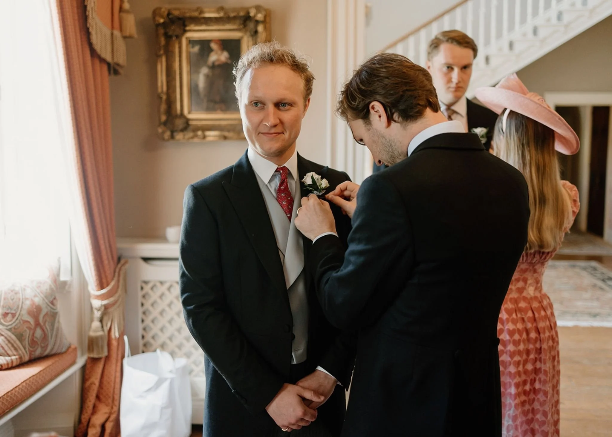 A man in a suit is being helped with his boutonniere at a formal event, with other attendees and elegant decor in the background.
