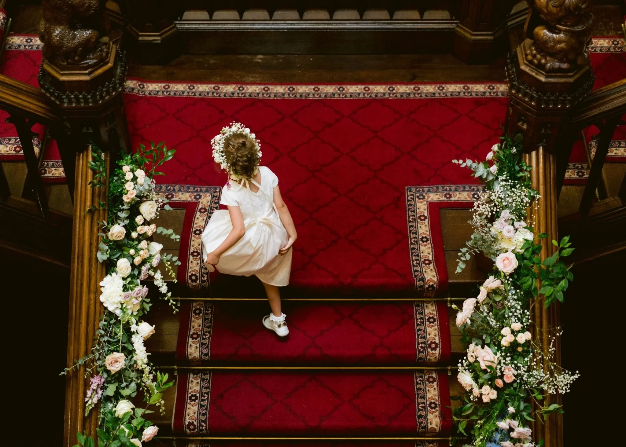 A young girl in a white dress with flowers in her hair stands on a red carpet at an ornate church altar decorated with flowers.