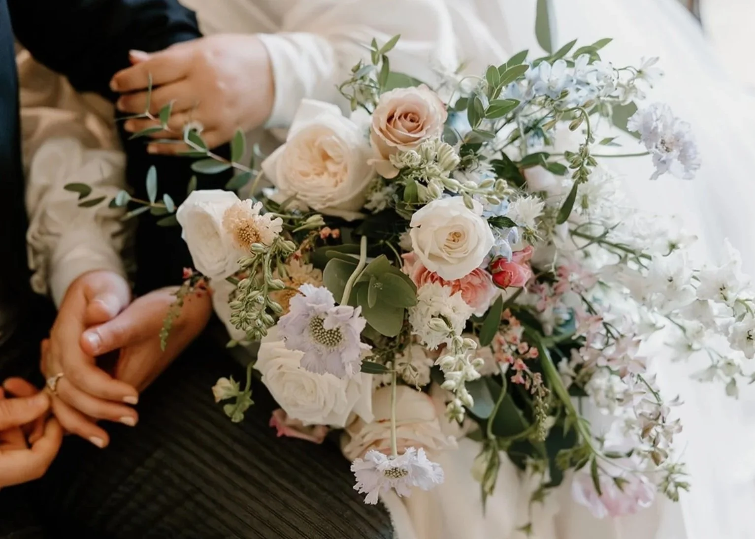 Close-up view of a wedding bouquet with white, blush pink, and pale purple flowers surrounded by green leaves, resting on a person's lap, with a wedding ring visible on the person's finger.