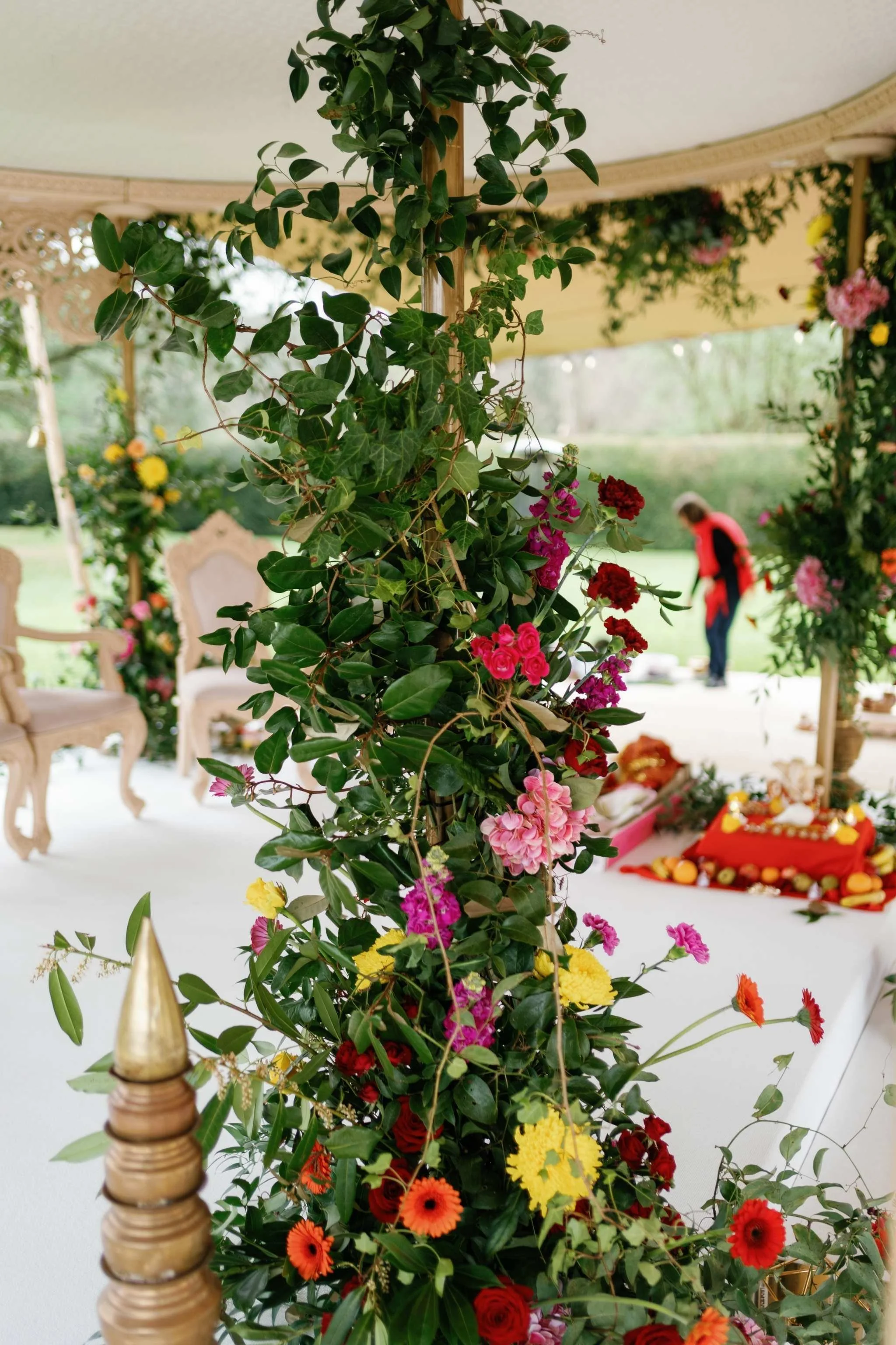 Close-up of a floral decoration with red, pink, orange, and yellow flowers intertwined with green leaves and ivy. In the background, chairs and a person are visible, suggesting a decorated outdoor or garden setting for an event.
